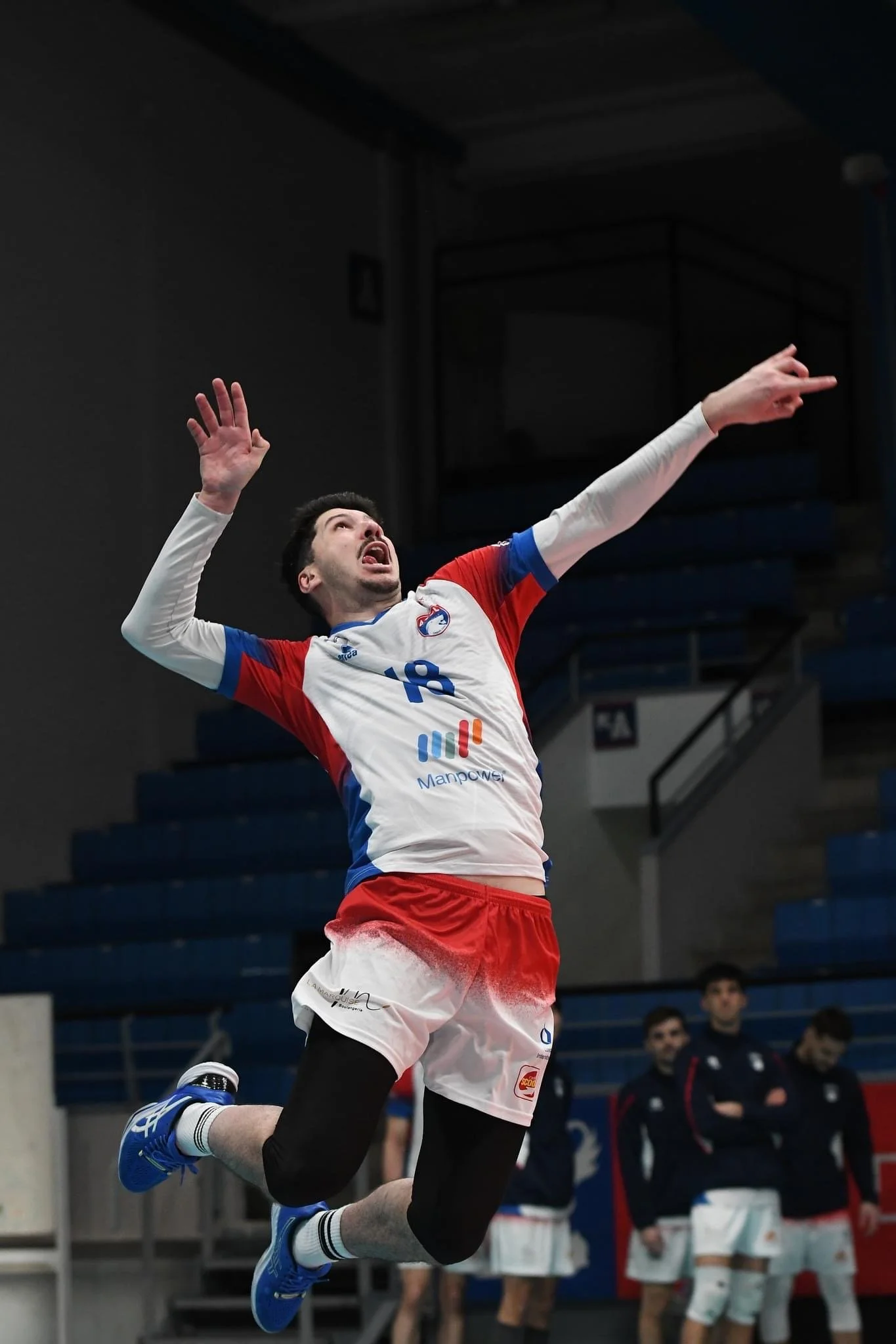 Un joueur de volleyball en plein saut lors d'un match, portant un maillot de volley, avec des coéquipiers en arrière-plan.