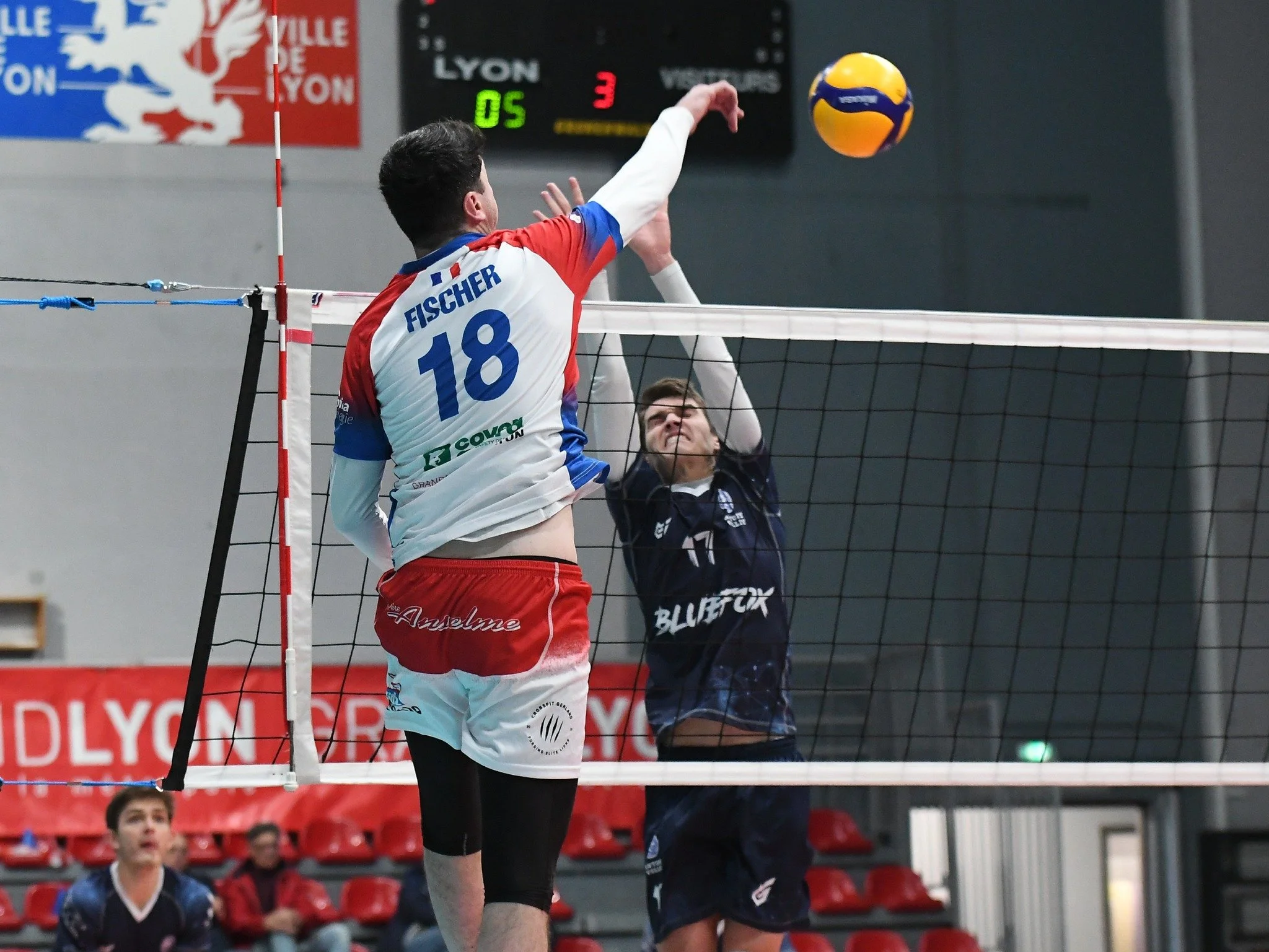 Deux joueurs de volleyball en action lors d'un match en salle. Le joueur en maillot blanc et rouge, numéro 18, est en train d'attaquer le ballon et le joueur en maillot noir de bloquer. Le fond montre un tableau de score et quelques spectateurs.