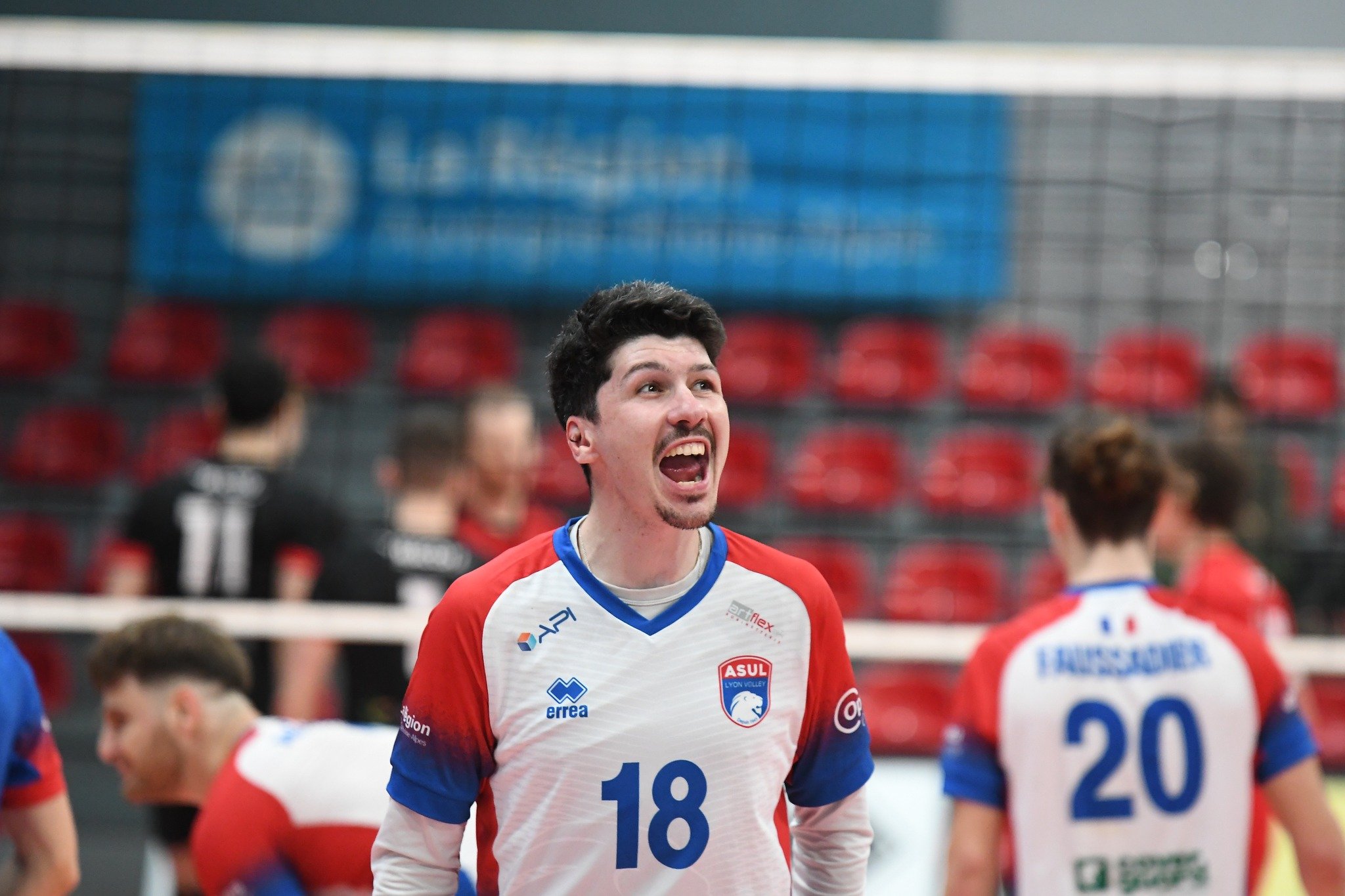 Un joueur de volley-ball en pleine célébration lors d'un match, portant un maillot blanc avec des détails en rouge et bleu, numéro 18, dans une salle sportive avec des spectateurs en arrière-plan.
