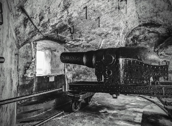 An old black cannon in a stone-walled tunnel or room, with a small window letting in natural light.