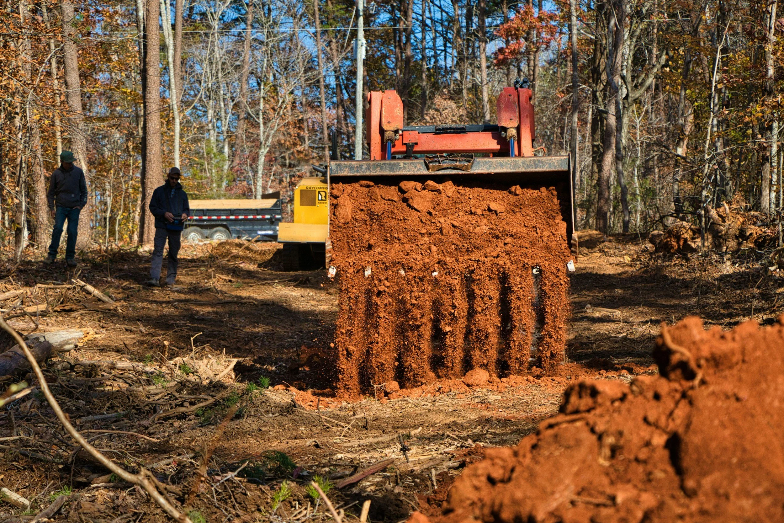 A large piece of construction equipment spewing reddish dirt onto the ground in a forested area during daytime.