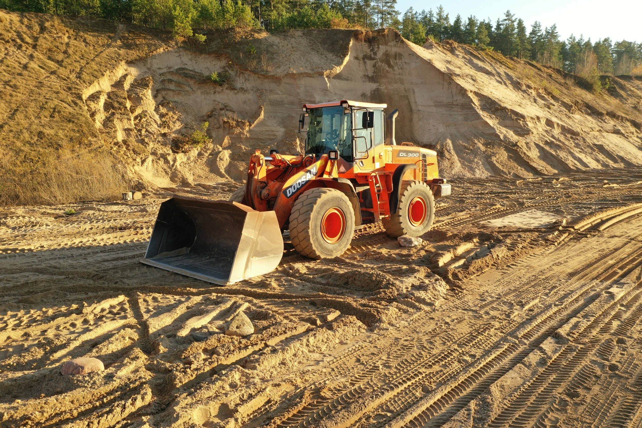 A construction site with a large orange Doosan DL300 wheel loader in the foreground, dirt and tire tracks on the ground, and a construction dirt wall with trees behind it in the background.