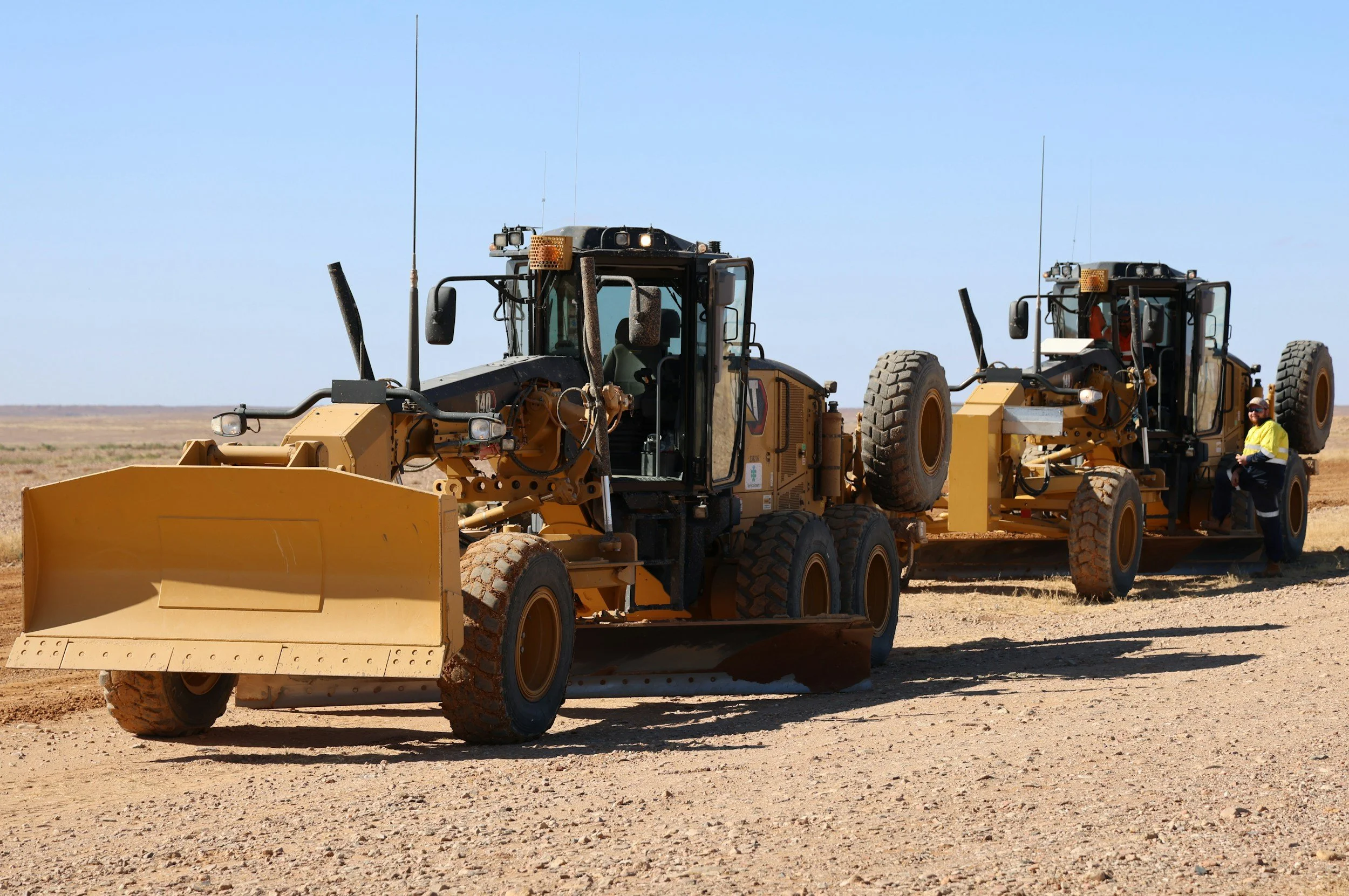 Two large yellow construction vehicles, bulldozers, working on a barren dirt field under a clear blue sky, with a worker standing beside one of the vehicles.