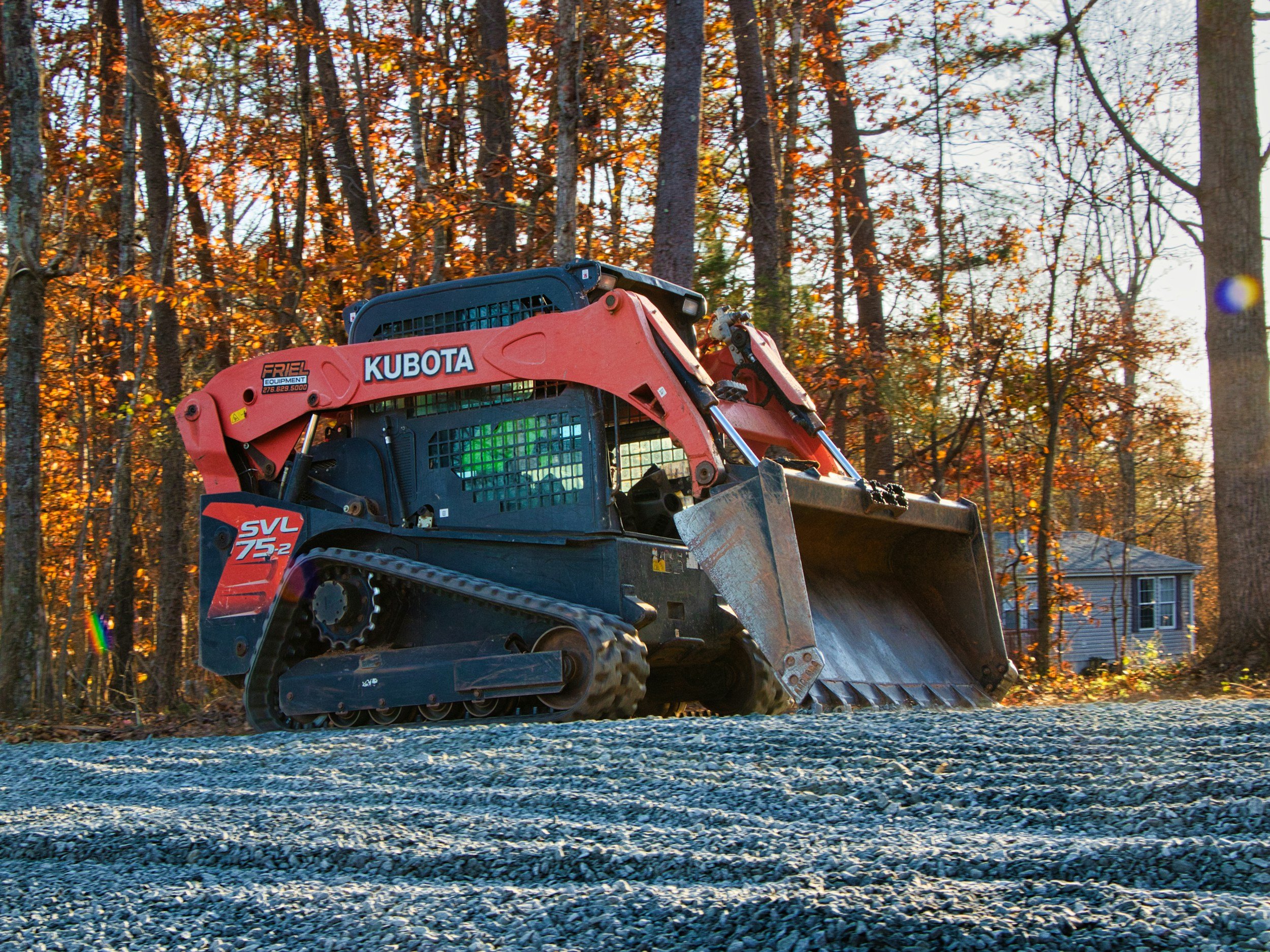 A construction vehicle, specifically a Kubota SVL 75.2 skid steer loader, working on a gravel surface in a wooded area during the fall season with trees showing autumn foliage in the background.
