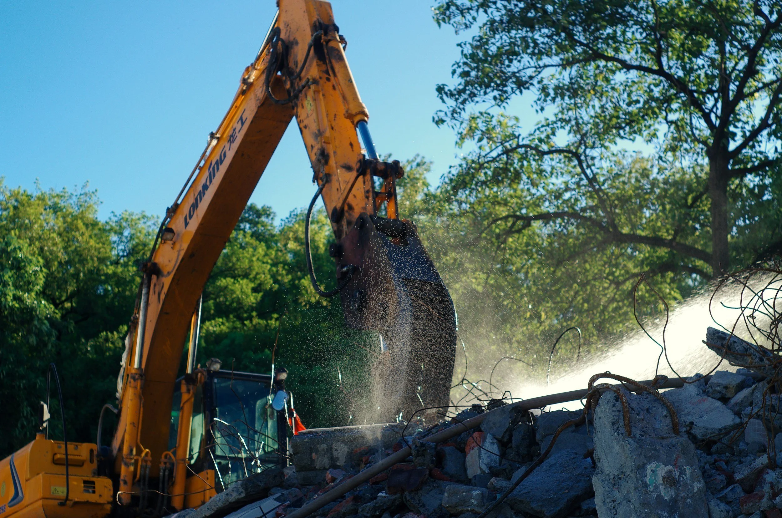 A construction excavator is demolishing concrete debris with a hydraulic hammer attachment, surrounded by trees and a clear blue sky.