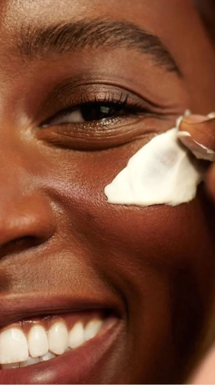 Pessoa sorrindo com maquiagem nos olhos, aplicando creme na bochecha