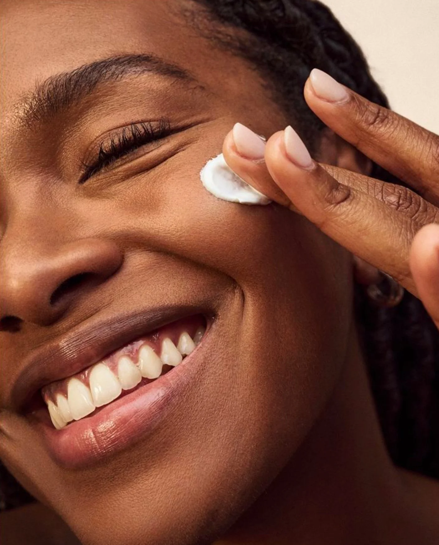 Pessoa sorrindo com creme de rosto sendo aplicando na bochecha.