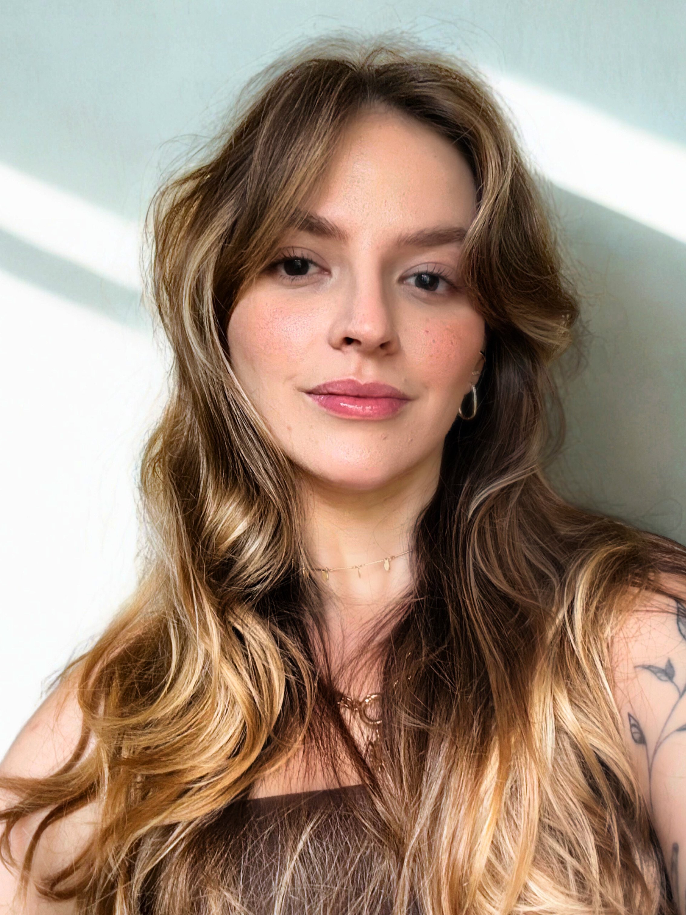 Retrato de uma mulher com cabelos longos e ondulados, tom de pele claro, usando brincos de argola, colar delicado e um vestido escuro, com fundo claro e luz suave.