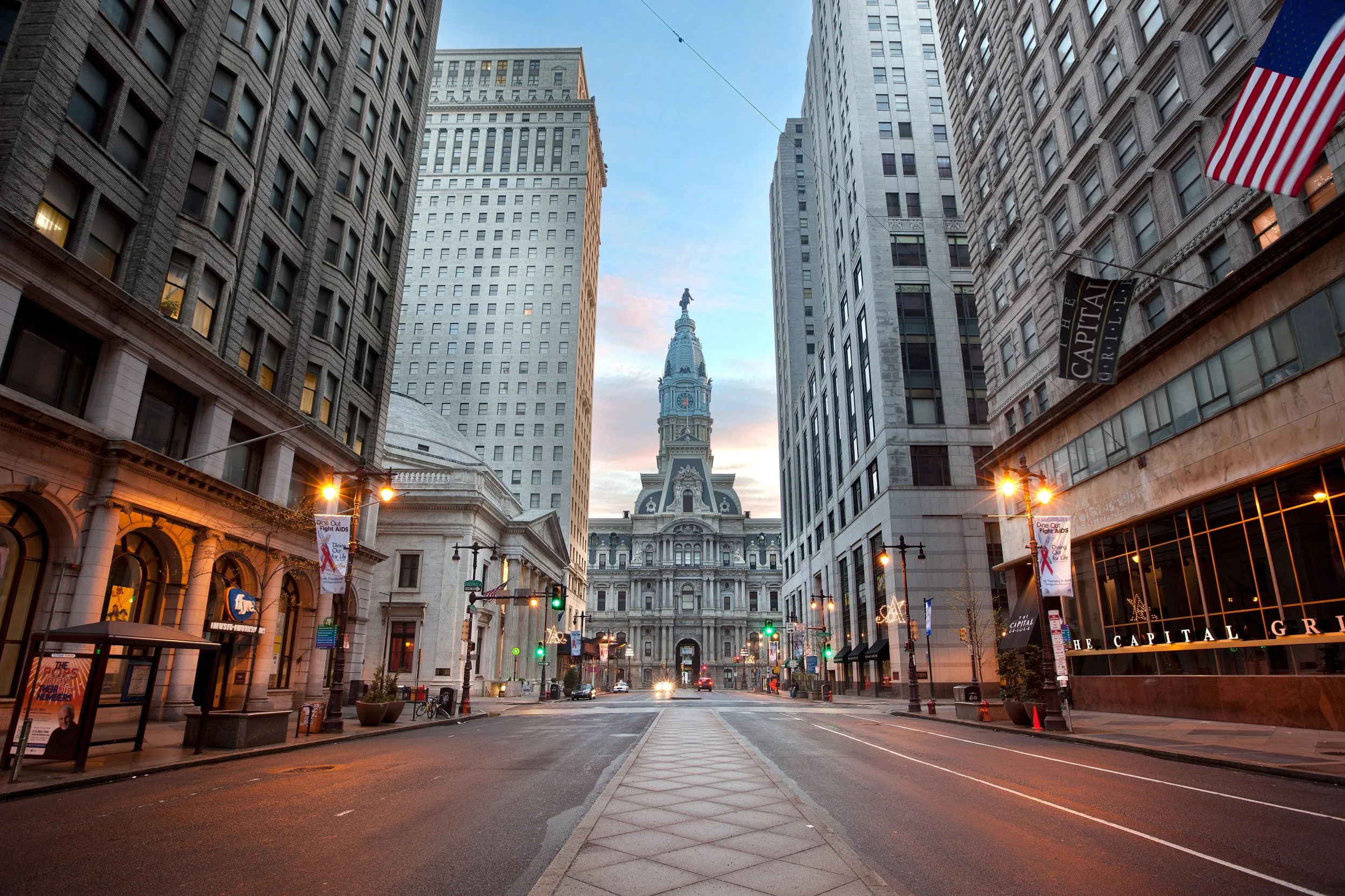 Empty city street in Philadelphia at dusk, with historic buildings and the Philadelphia City Hall with a statue on top, traffic lights and banners visible.