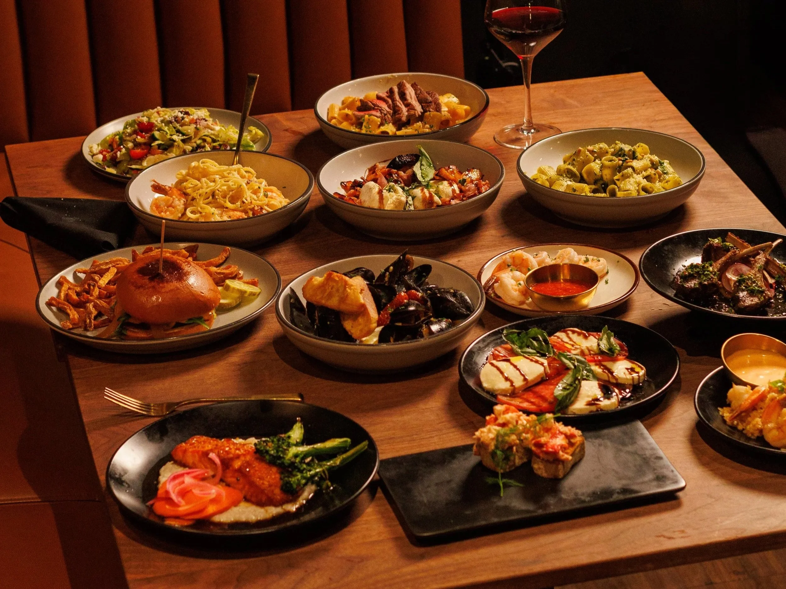 A table filled with various dishes including pasta, salad, seafood, and meat, with a glass of red wine.