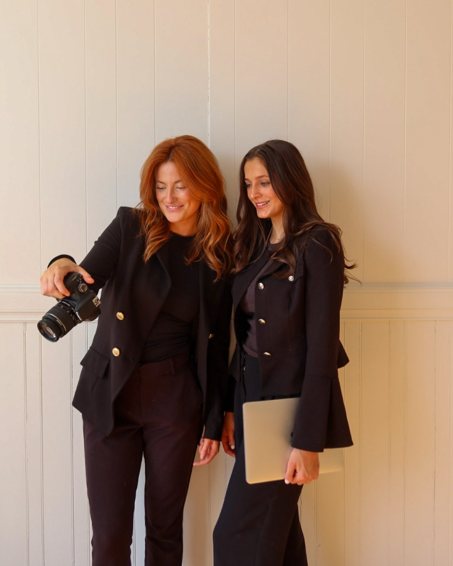 Two women dressed in black suits standing close together, smiling, one holding a camera and the other holding a laptop, against a light-colored wooden wall.