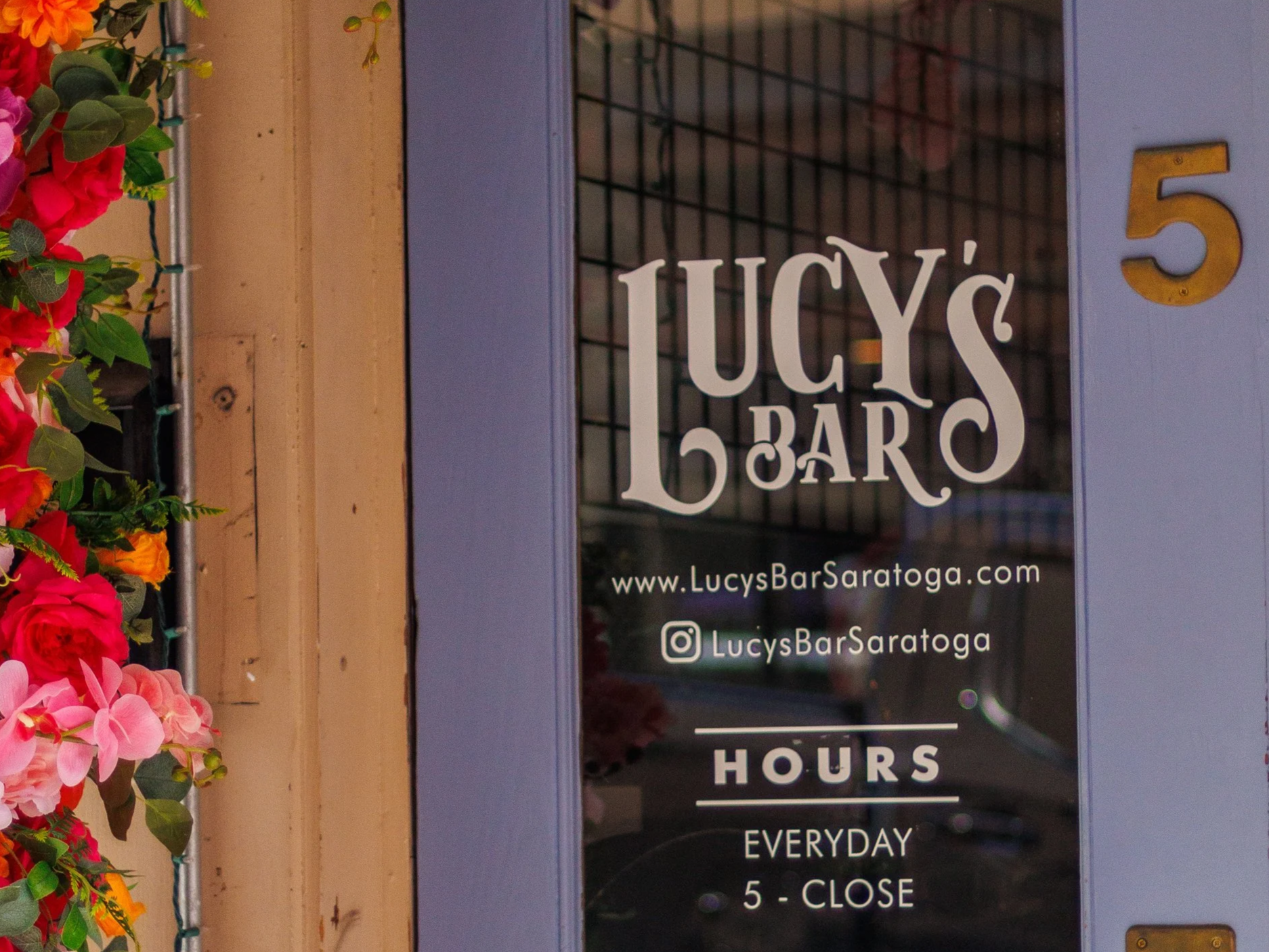 A sign for Lucy’s Bar with the address 5, surrounded by a flower arrangement on the left side. The sign includes the website www.LucysBarSaratoga.com and social media handle @LucysBarSaratoga. It states the opening hours as every day from 5 PM until close.