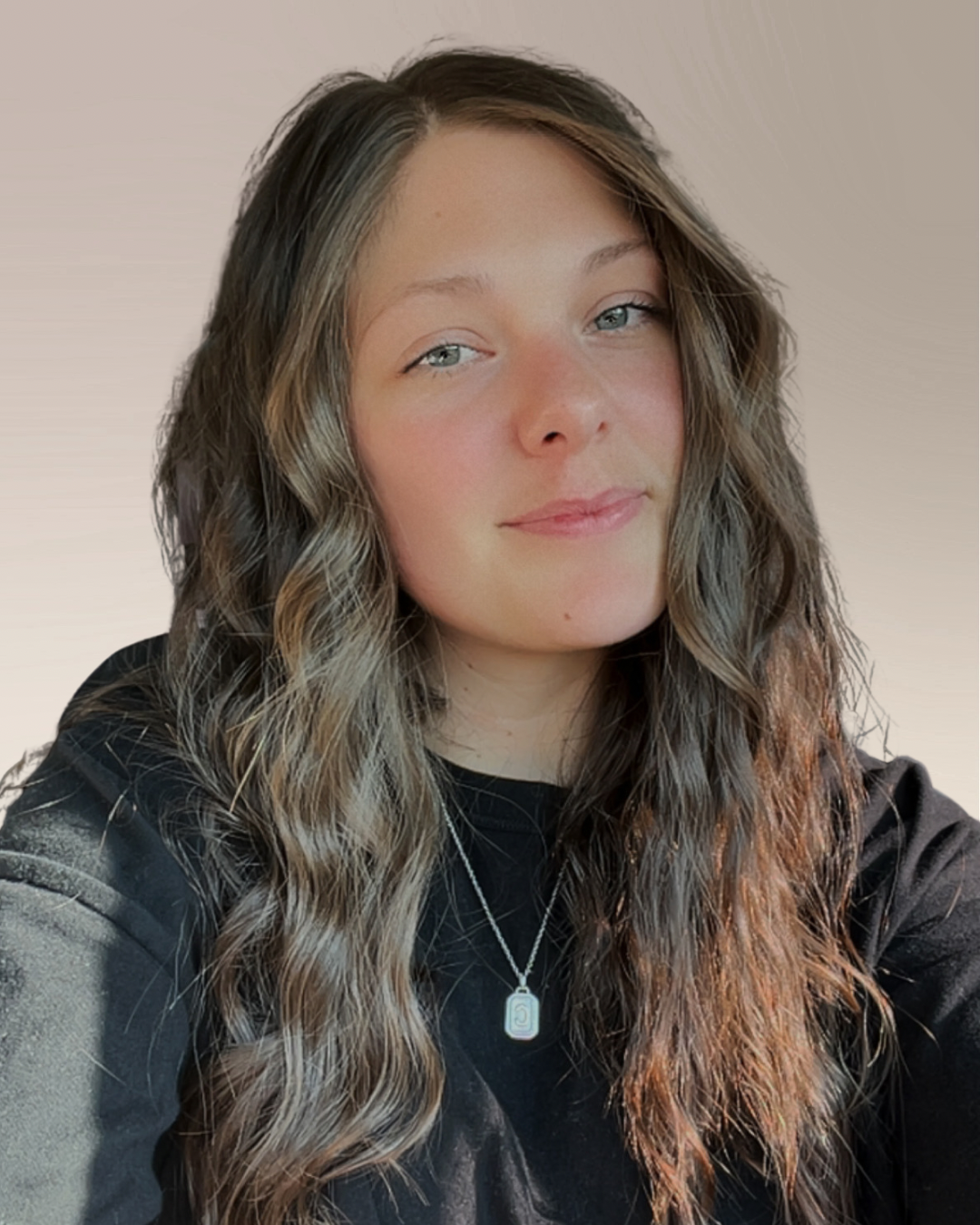 A young woman with wavy brown hair and fair skin, wearing a black top and a silver necklace with a pendant, smiling softly.