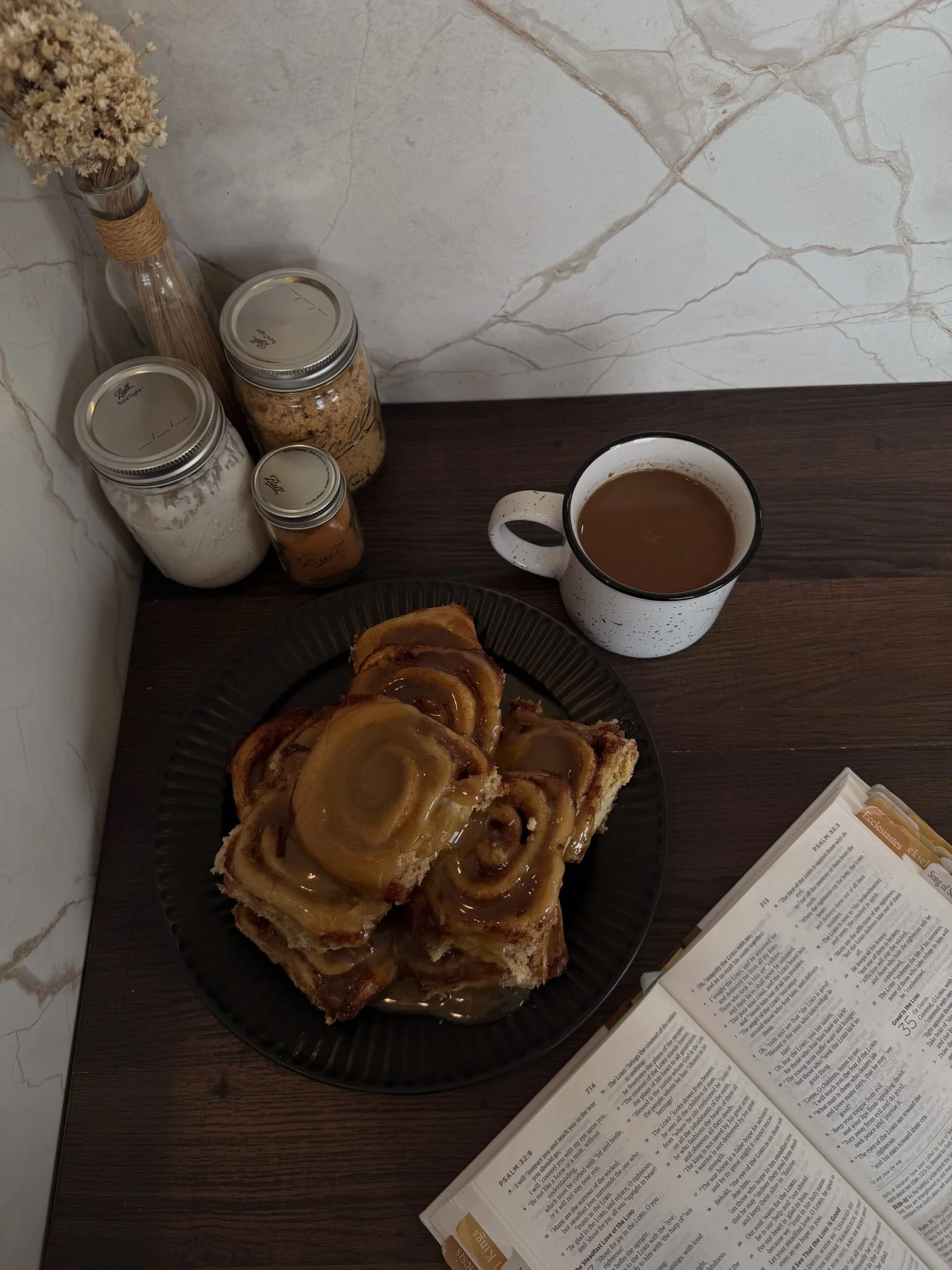 Plate of cinnamon rolls with caramel glaze next to a cup of coffee with cream, alongside jars of sugar and spices, an open Bible, and a vase with dried flowers on a wooden table against a marble wall.