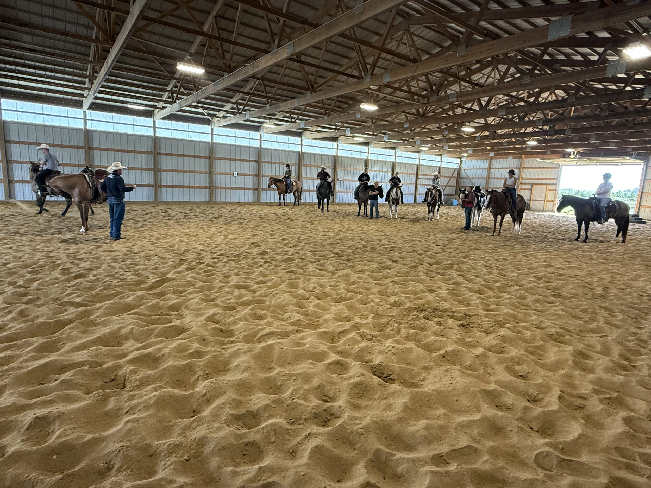 People riding horses inside a large indoor arena with sandy footing and a high wooden ceiling.