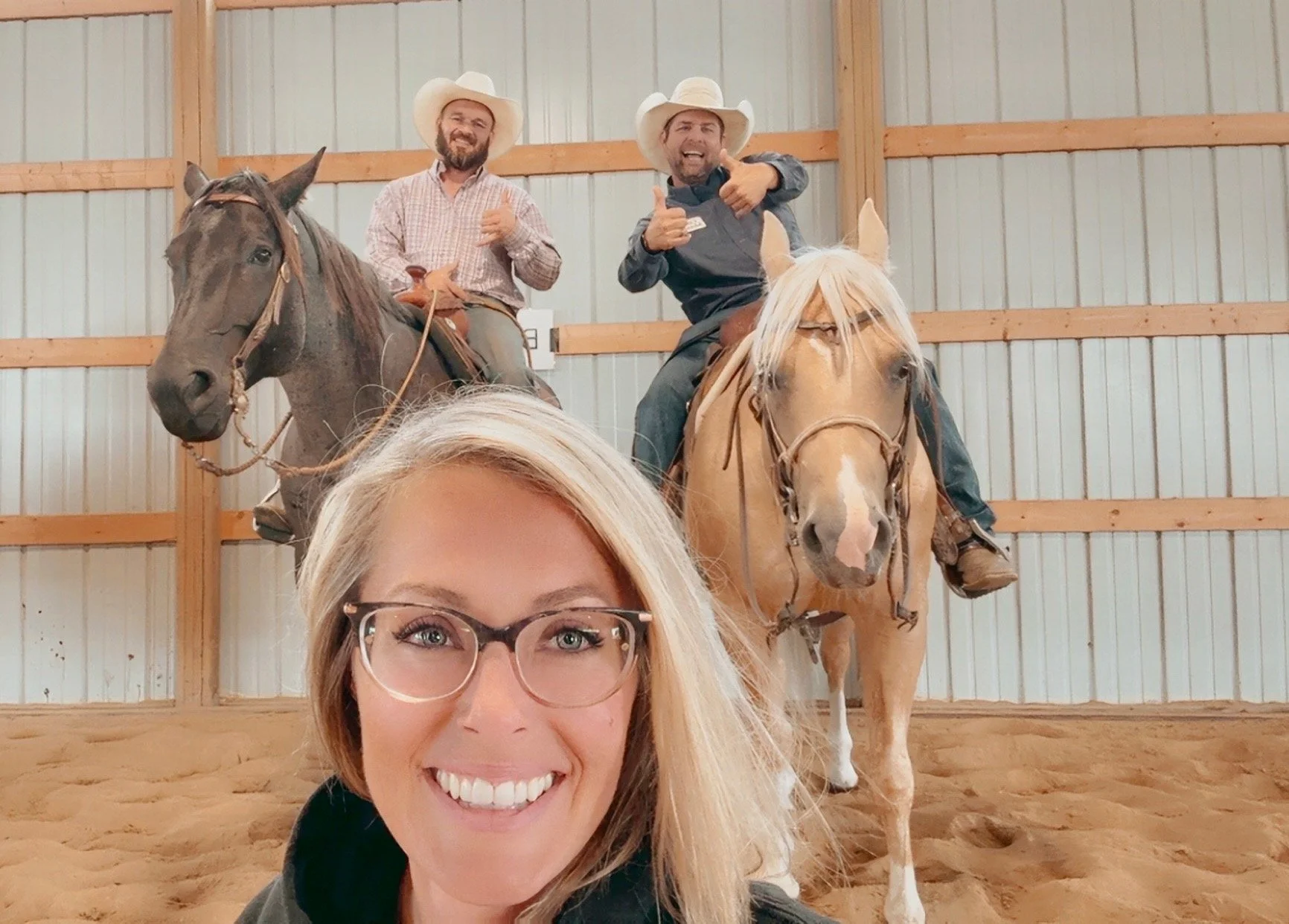 A woman wearing glasses takes a selfie in front of two men riding horses inside a wooden indoor arena with sandy ground. The men are wearing cowboy hats and are smiling and giving thumbs-up.