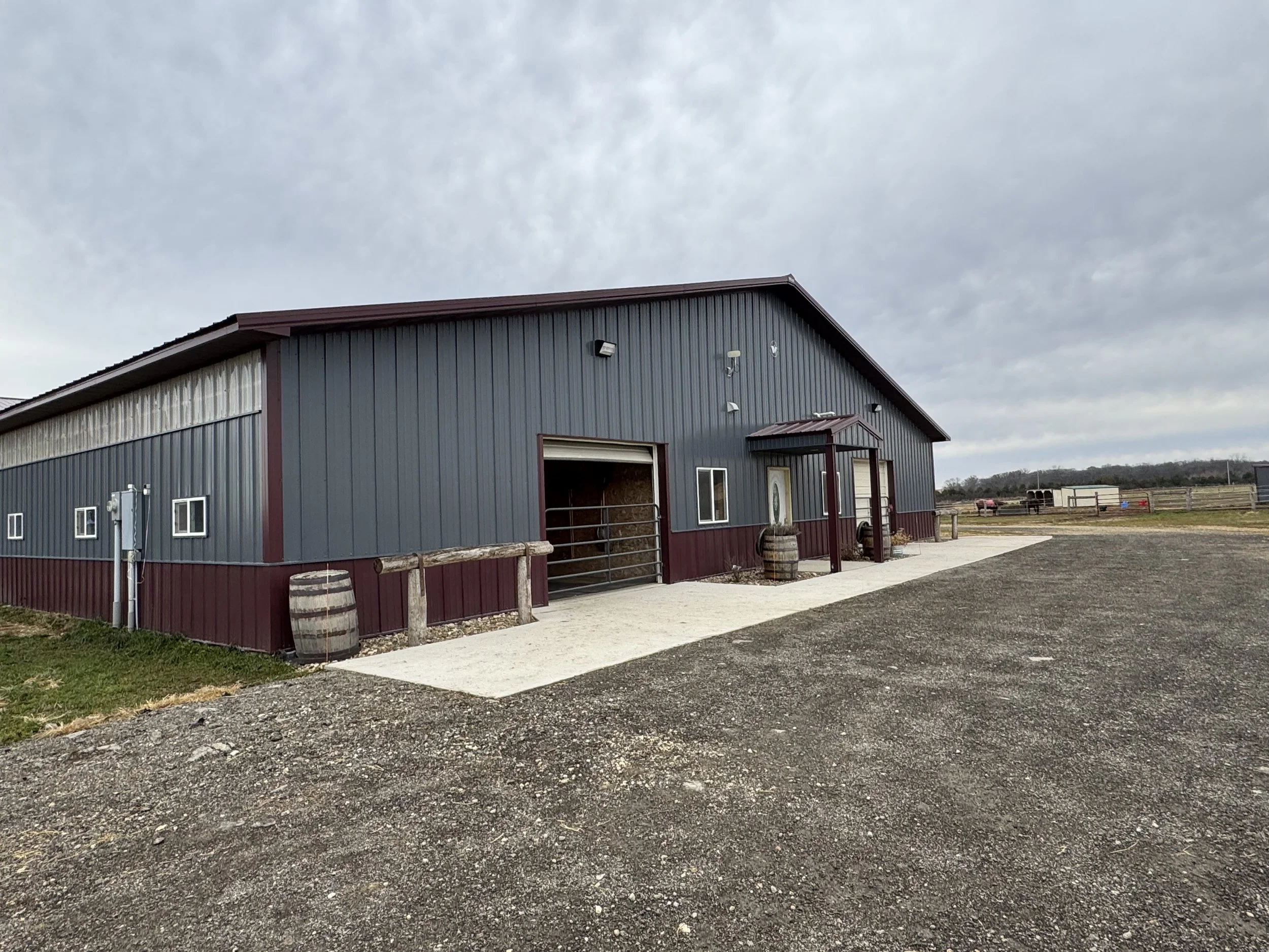 A large metal barn with burgundy trim, two small windows, and a small entrance door under a small porch in a rural area. There are several barrels and a wooden fence in front and a gravel parking area.