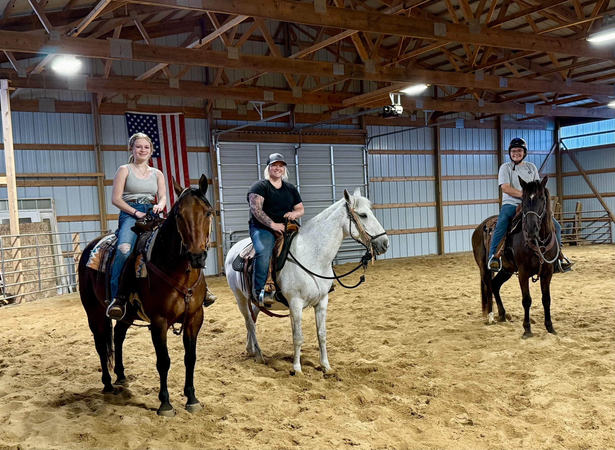 Three women riding horses inside an indoor riding arena, with American flag hanging on the wall behind them.