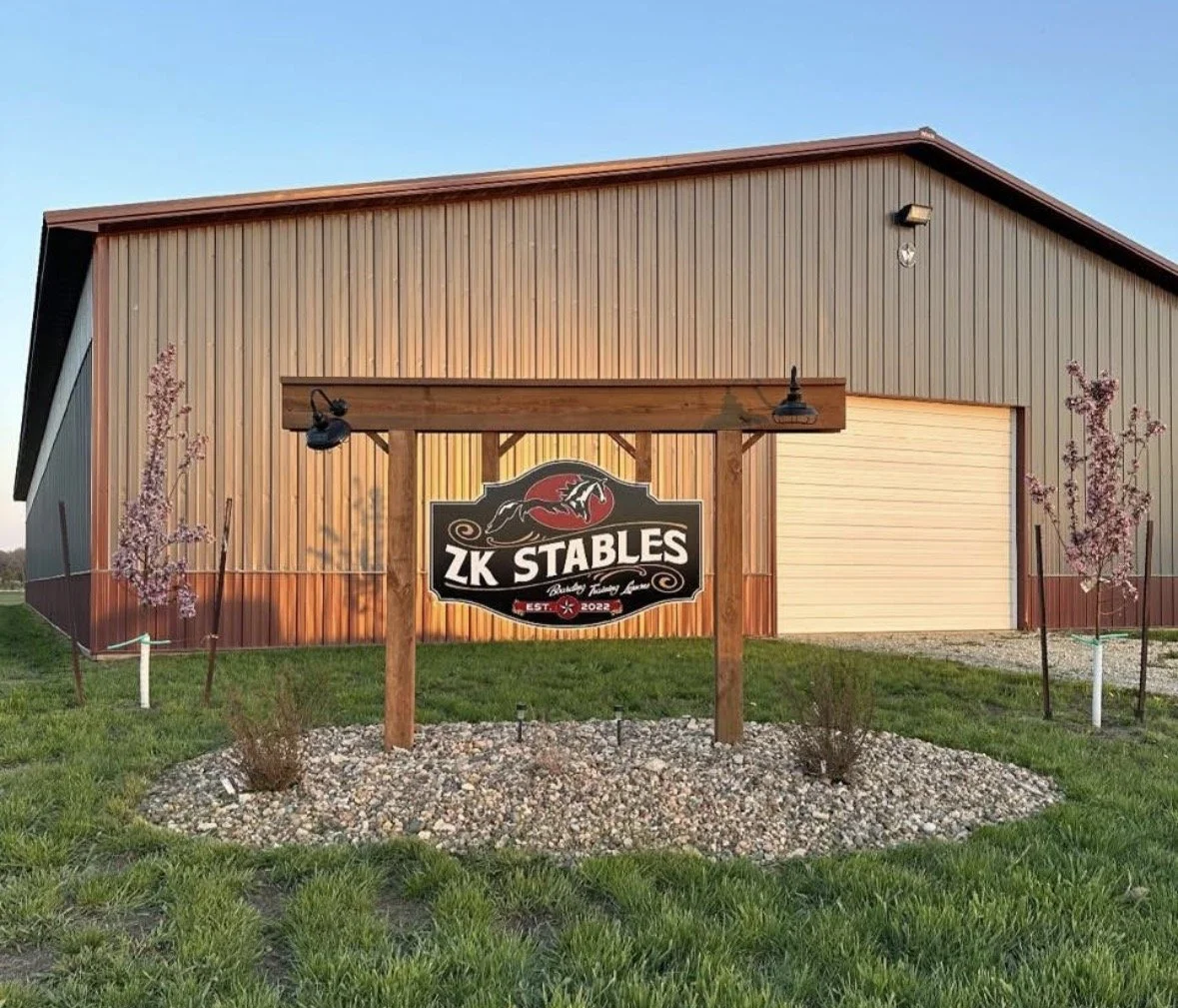 A large metal building with a sliding door and a wooden sign in front that reads 'ZK Stables'. The sign features a horse logo and the year 2022. There are pink flowering trees on either side of the sign, with a gravel bed and green grass surrounding it.