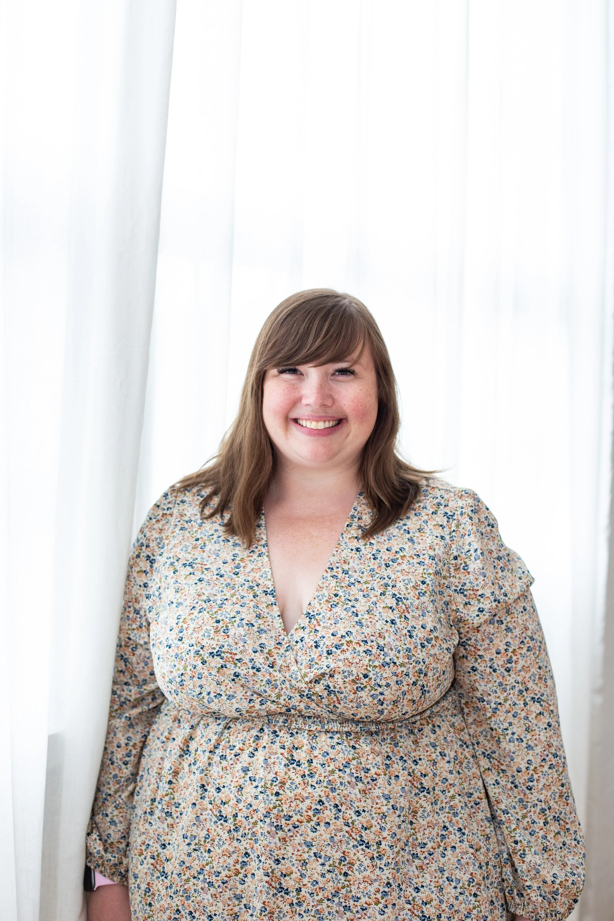 Kristina Bartold is  wearing a floral dress and standing in front of white curtains.