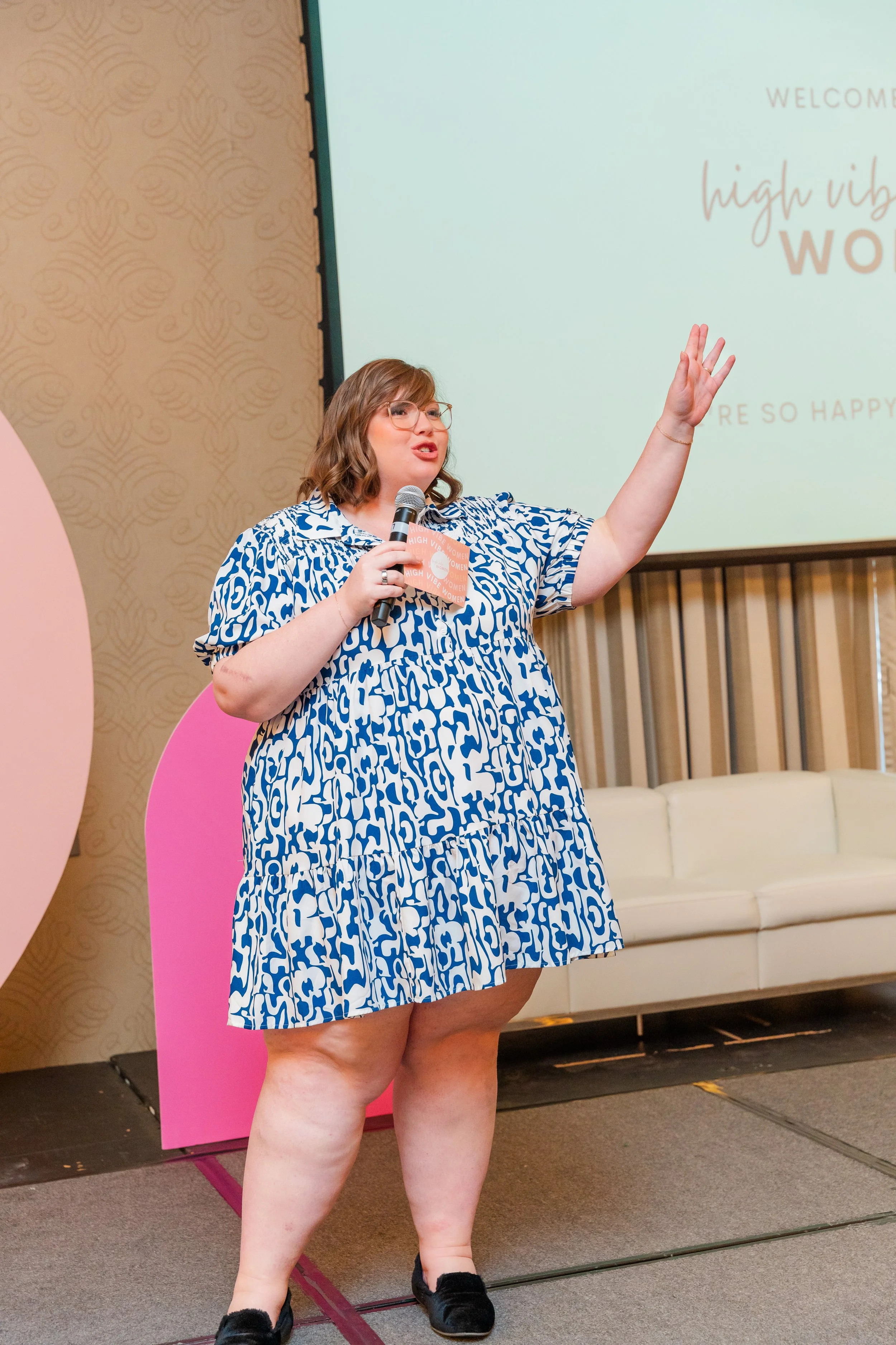 Kristina Bartold stands on stage holding a microphone in her right hand and raising her left hand. Behind her, a large screen displays a welcoming message with the words "high vibe woman" and "you're so happy," along with a pink and beige backdrop.