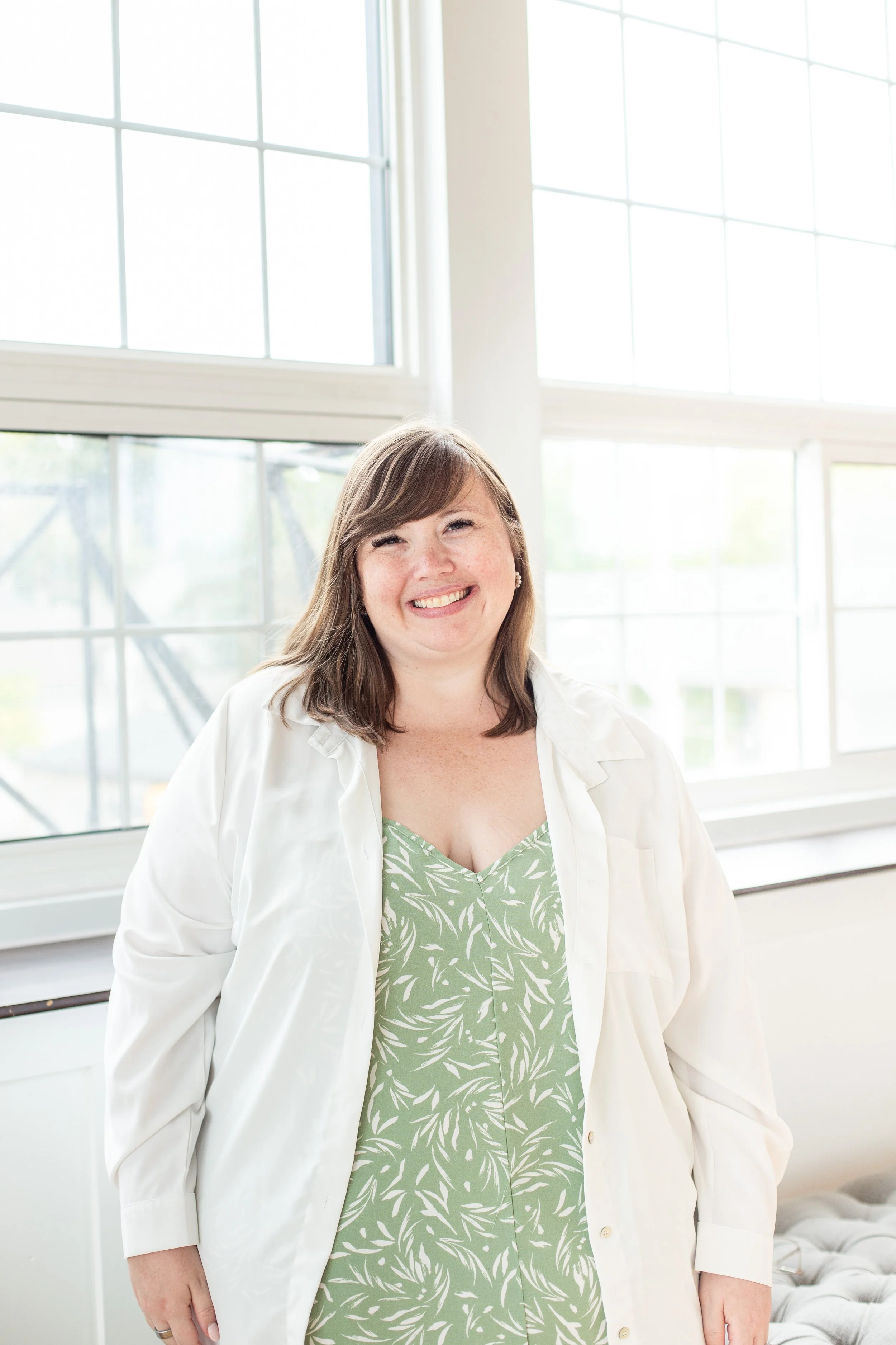 Kristina Bartold is standing indoors near large windows, smiling, wearing a light-colored jacket over a green dress with a white leaf pattern.