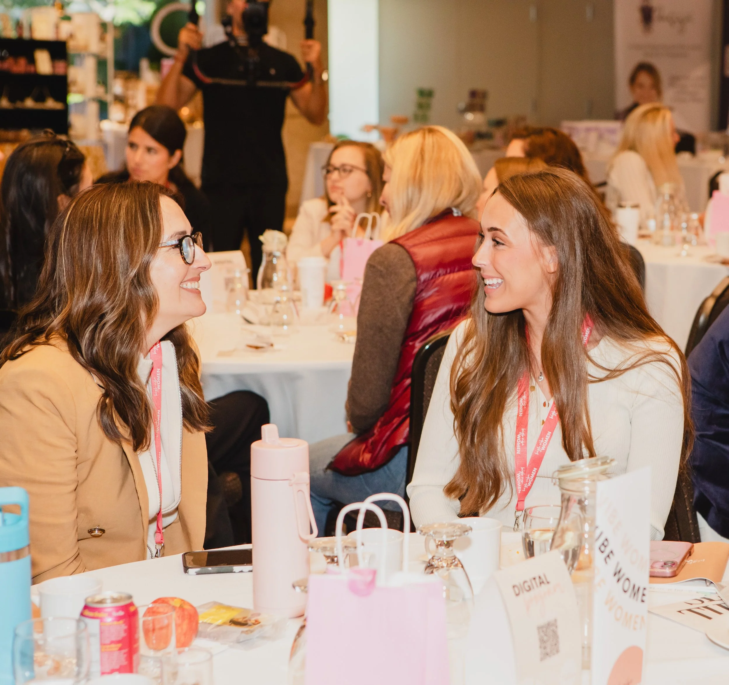 Two women smiling and engaging in conversation at a social event or conference, seated at a round table with drinks, pink gift bags, and event brochures.