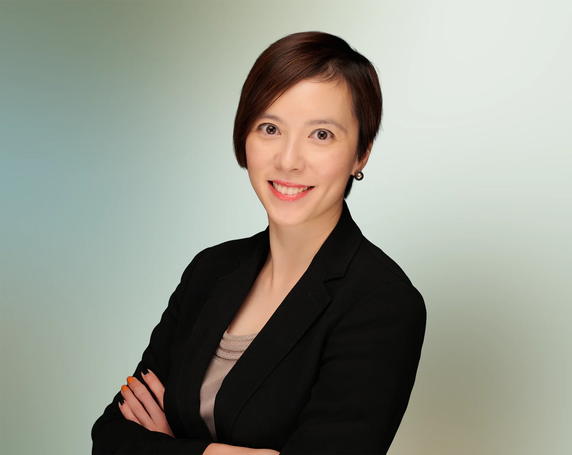 Professional woman with short dark hair, wearing a black blazer and a beige top, smiling with crossed arms, against a gradient background.
