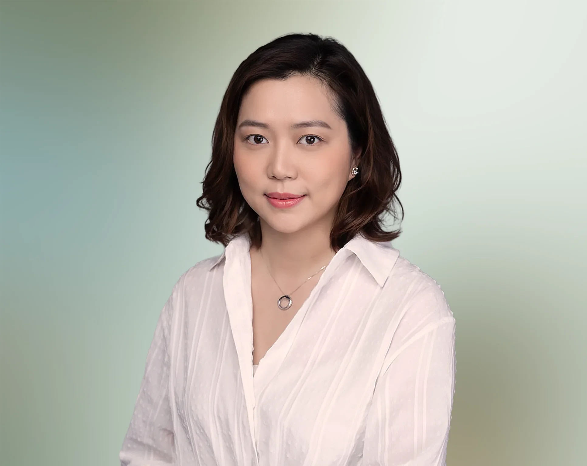 A young woman with shoulder-length dark hair, wearing a white blouse and a necklace, smiling softly against a blurred green background.