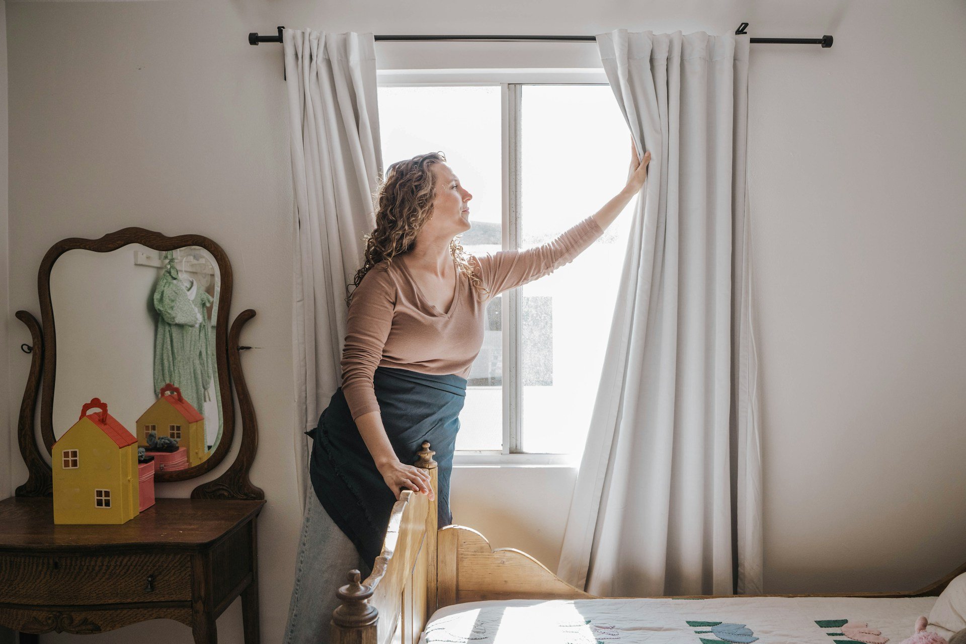 A woman with curly hair pulls back white curtains in a brightly lit bedroom, standing beside a wooden bed with a blanket. Behind her, a mirror on a small wooden dresser shows two yellow house-shaped decor items, a pink container, and some bottles. The room has white walls and a large window.