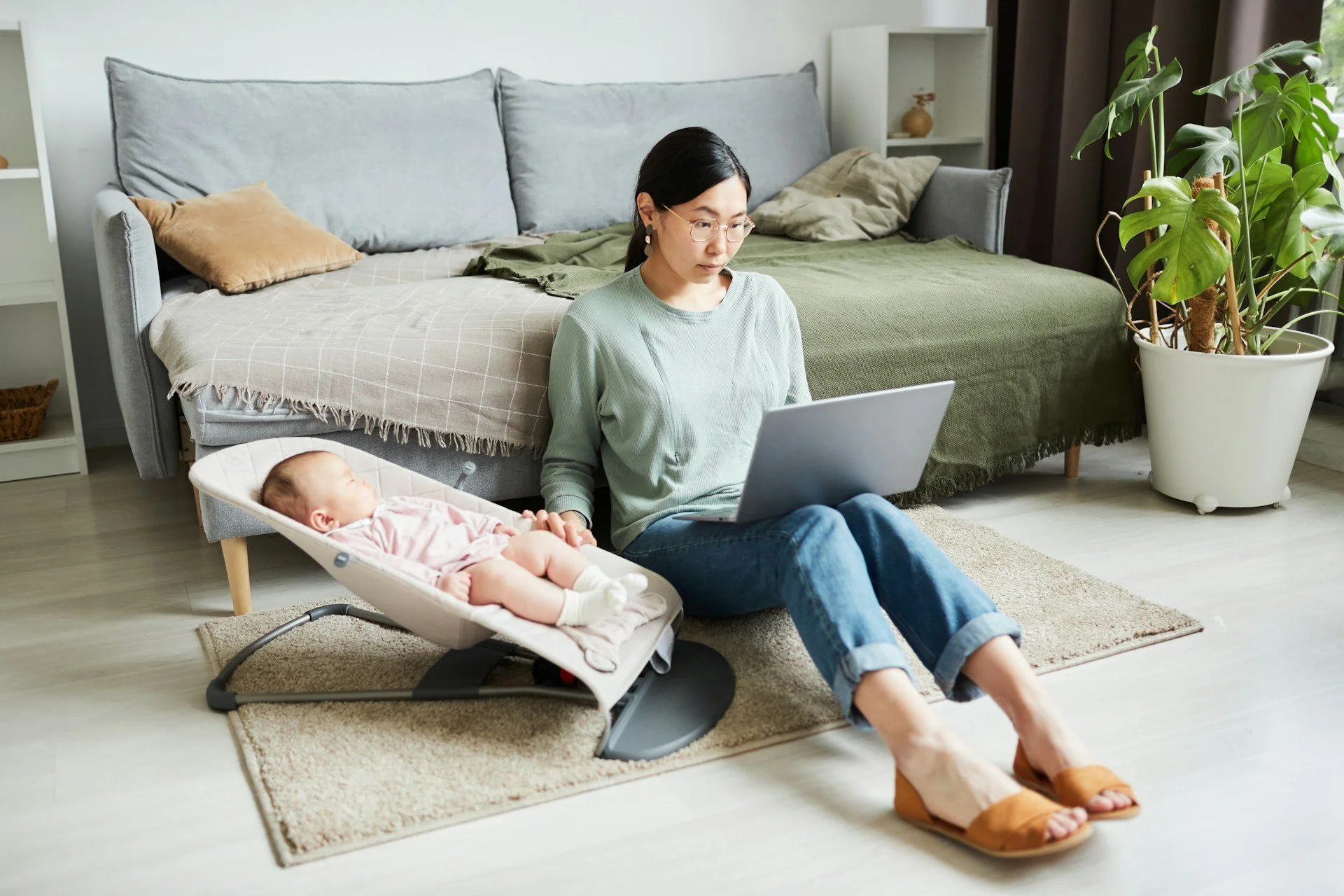 Woman sitting on the floor with a laptop, holding hands with a sleeping baby in a bassinet, in a bedroom with a bed and large potted plant.