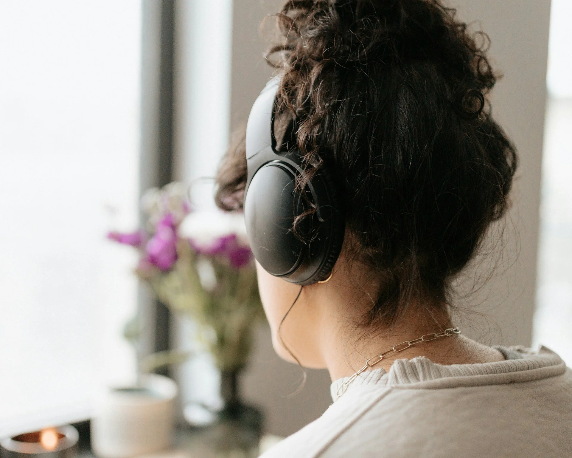 A person with curly hair wearing large headphones, looking at a computer or screen near a window with a vase of purple flowers in the background.
