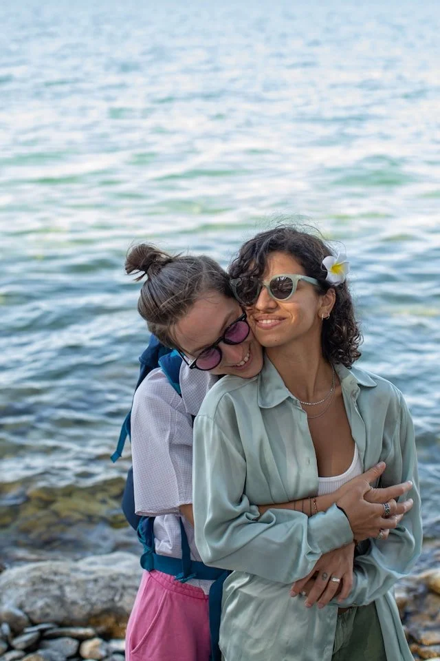 Two women embracing by the water, smiling, with the ocean in the background. They are wearing sunglasses and casual clothing, with one woman wearing a white top and the other a light green jacket.