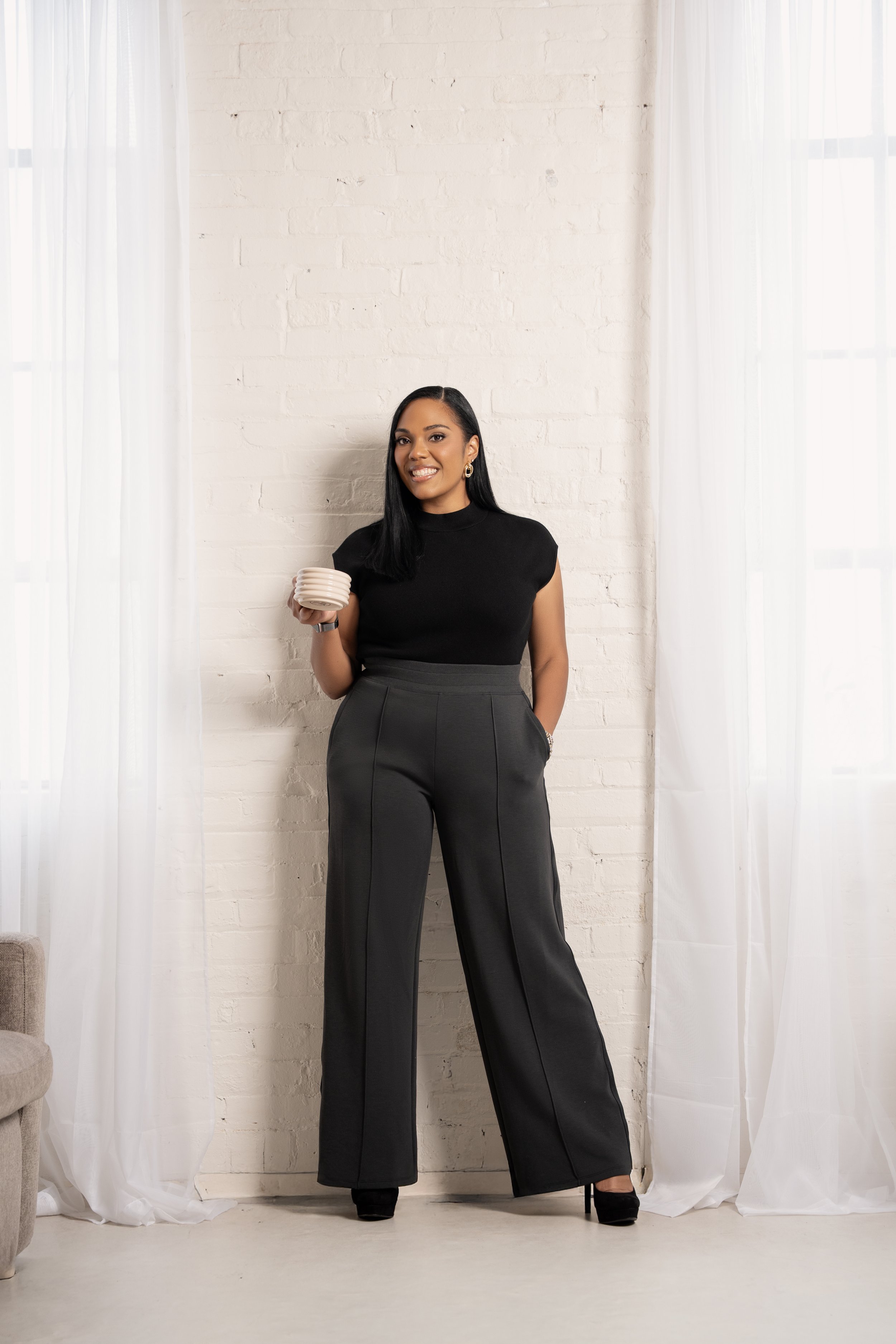A woman with medium skin tone, long black hair, wearing a black top and wide-leg gray pants, standing with one hand in her pocket and holding a mug in the other, in front of a white brick wall with sheer curtains on each side.