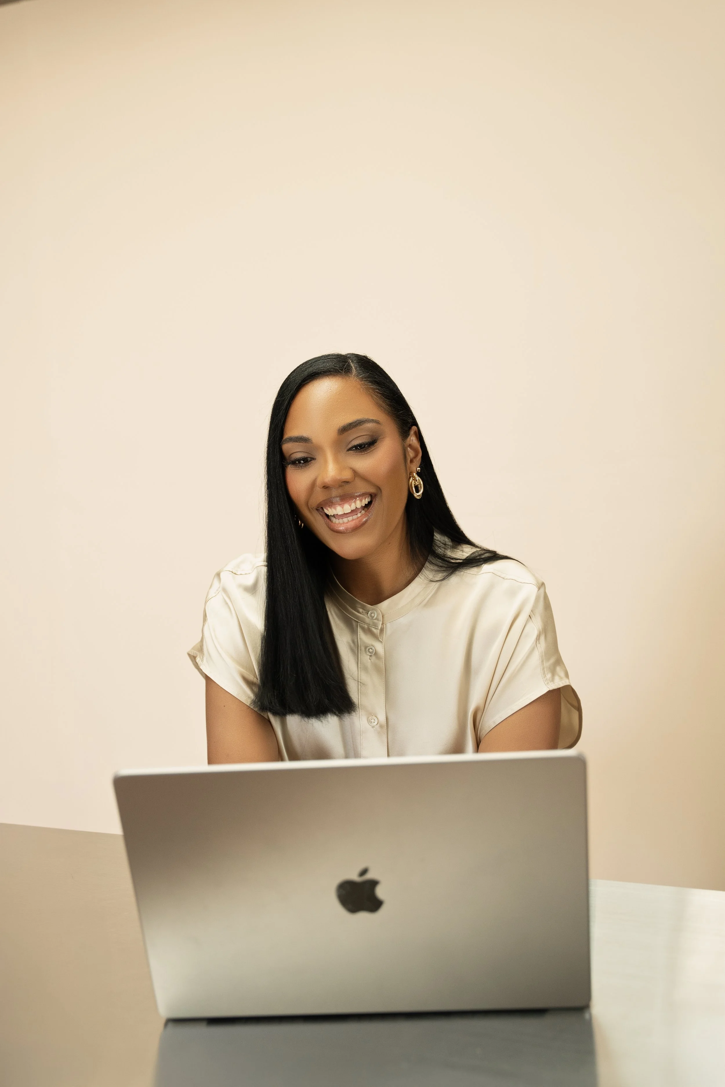 A woman with long black hair wearing a light-colored silk blouse, smiling and looking at a silver MacBook in front of her.