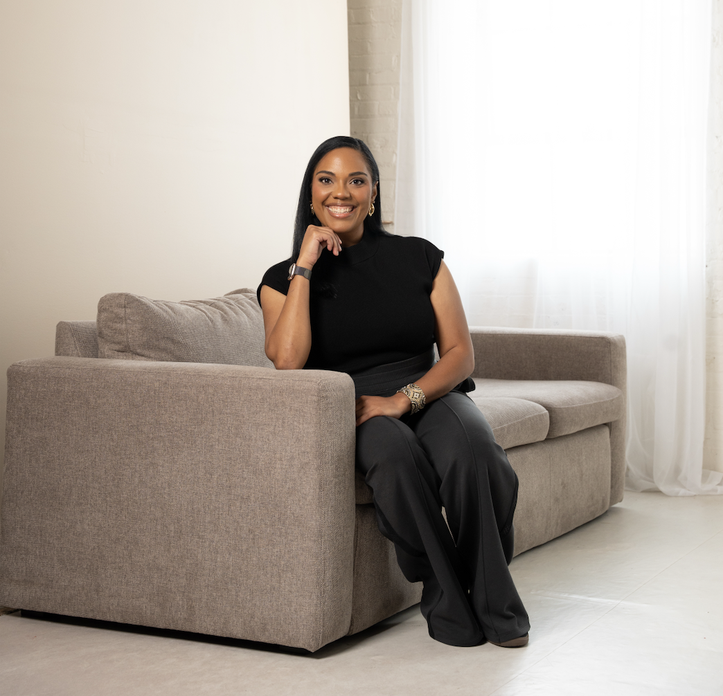 A woman sitting on a beige sofa, smiling, wearing a black top and black pants, in a room with white curtains and a white brick wall.
