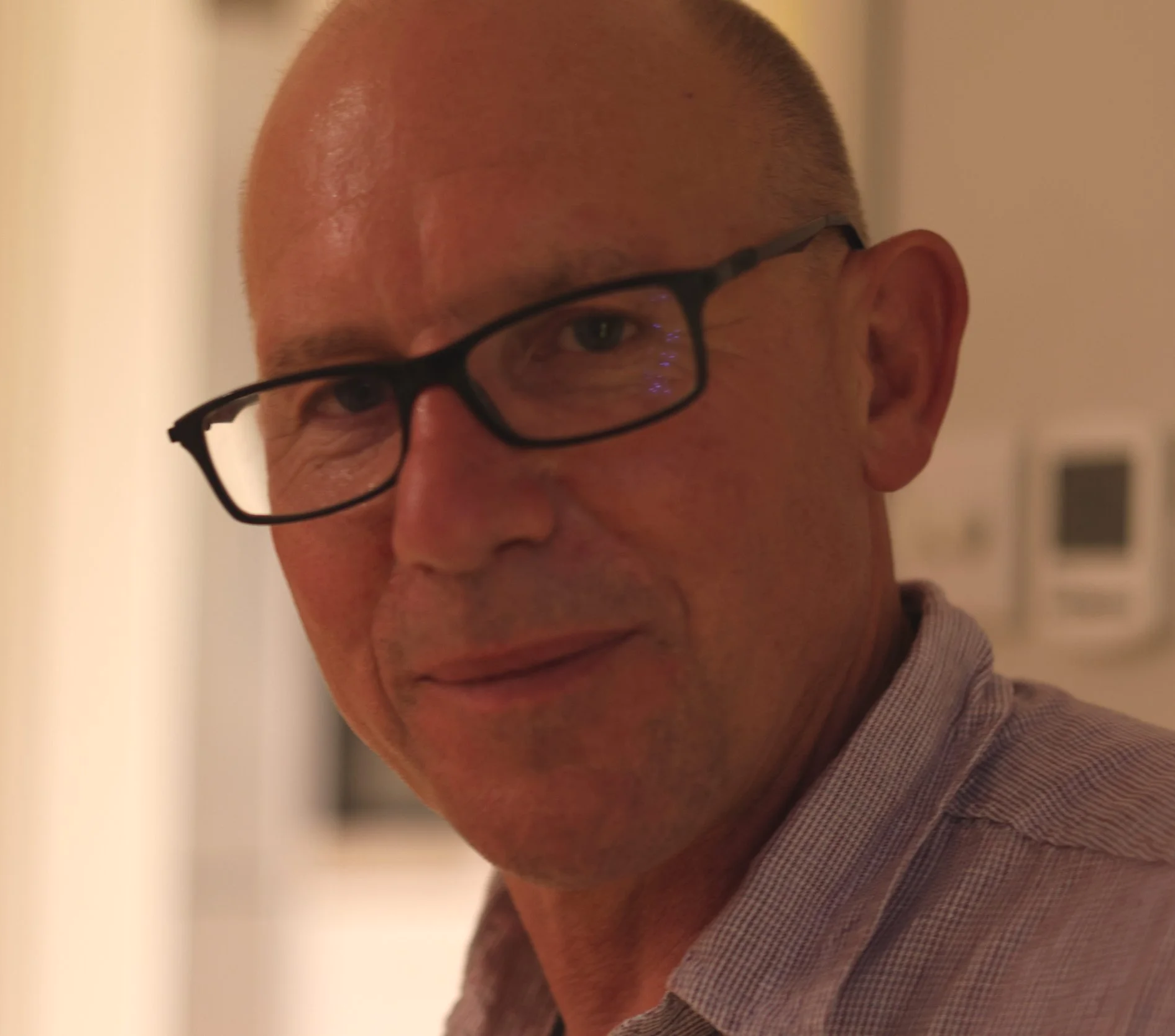 A close-up of a man with short hair, wearing black glasses, smiling slightly, dressed in a checkered shirt, with a blurred background.