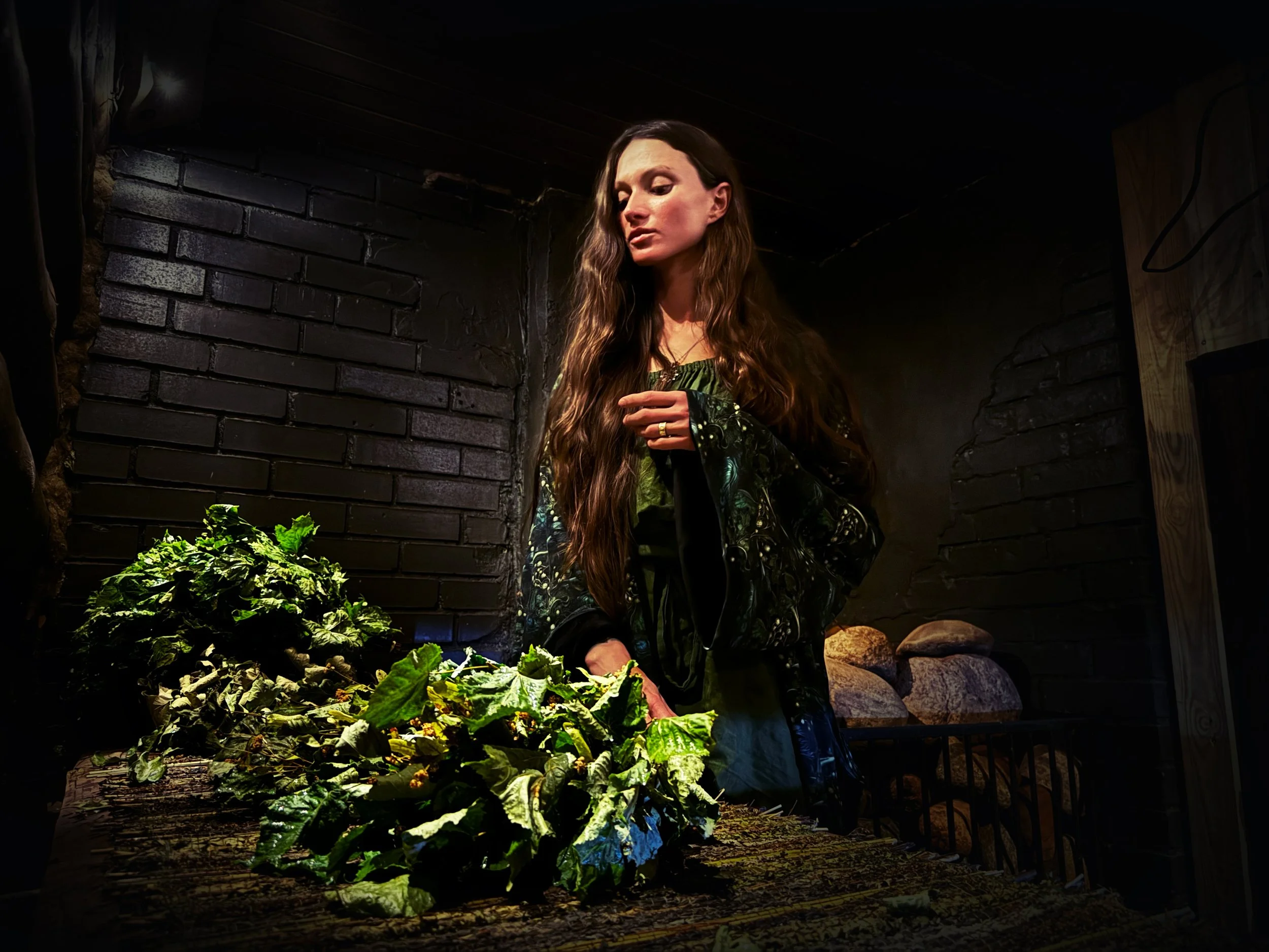 A woman with long, wavy brown hair standing in a dark room with black brick walls, surrounded by leafy greens on a rustic wooden table.