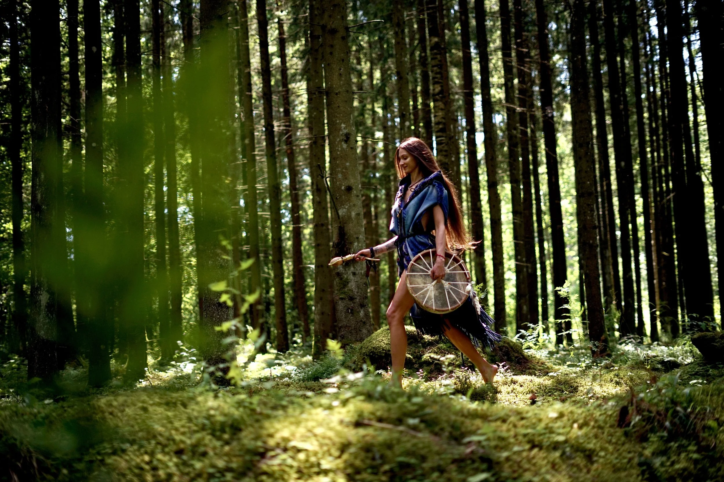 A woman dressed in tribal attire, holding a shield and a spear, walking barefoot through a dense forest with tall trees and green moss on the ground.