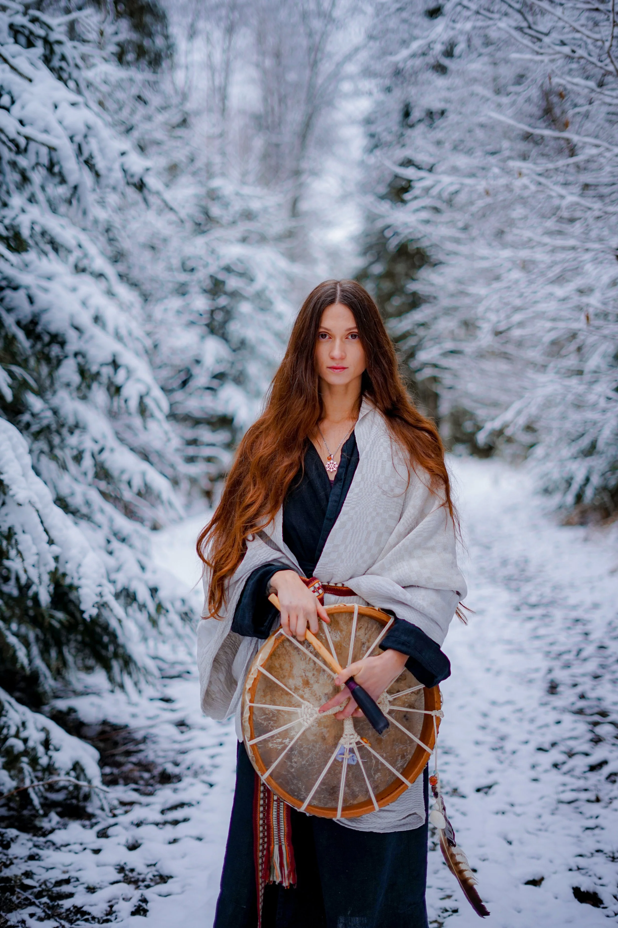 A woman with long, wavy, brown hair standing in a snow-covered forest, holding a large traditional drum.