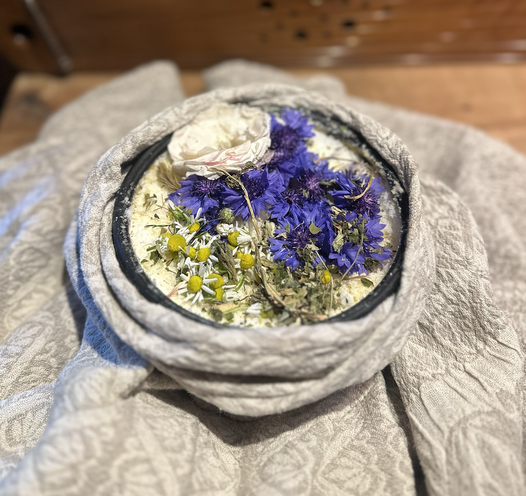 A close-up of a container filled with a mixture of dried flowers, including purple, white, and yellow blooms, placed on a cloth surface.