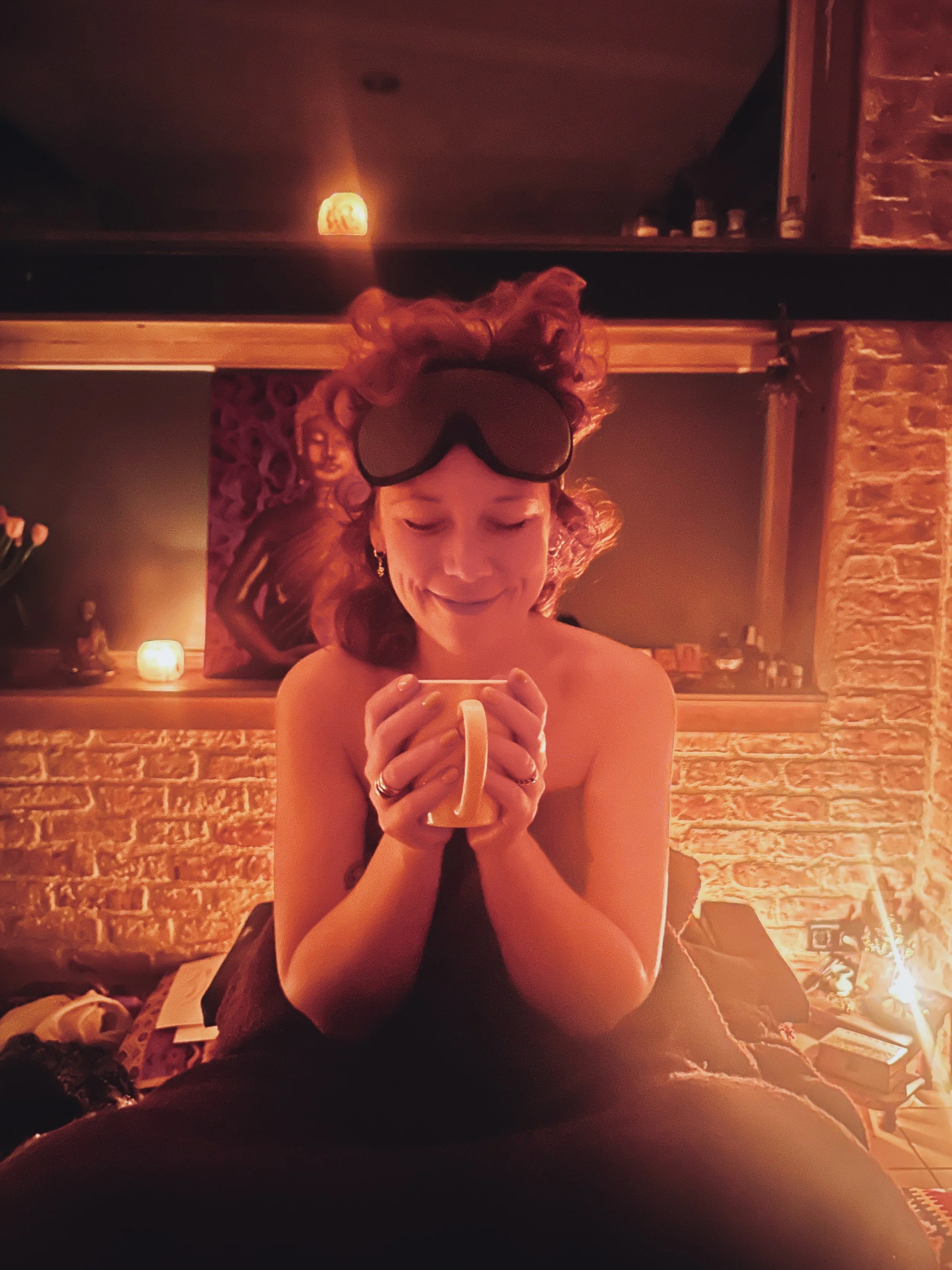 A woman sitting in a dimly-lit room, holding a mug, with her eyes closed and smiling. She has curly hair with a large bow or headband, and is wearing a strapless top or dress.