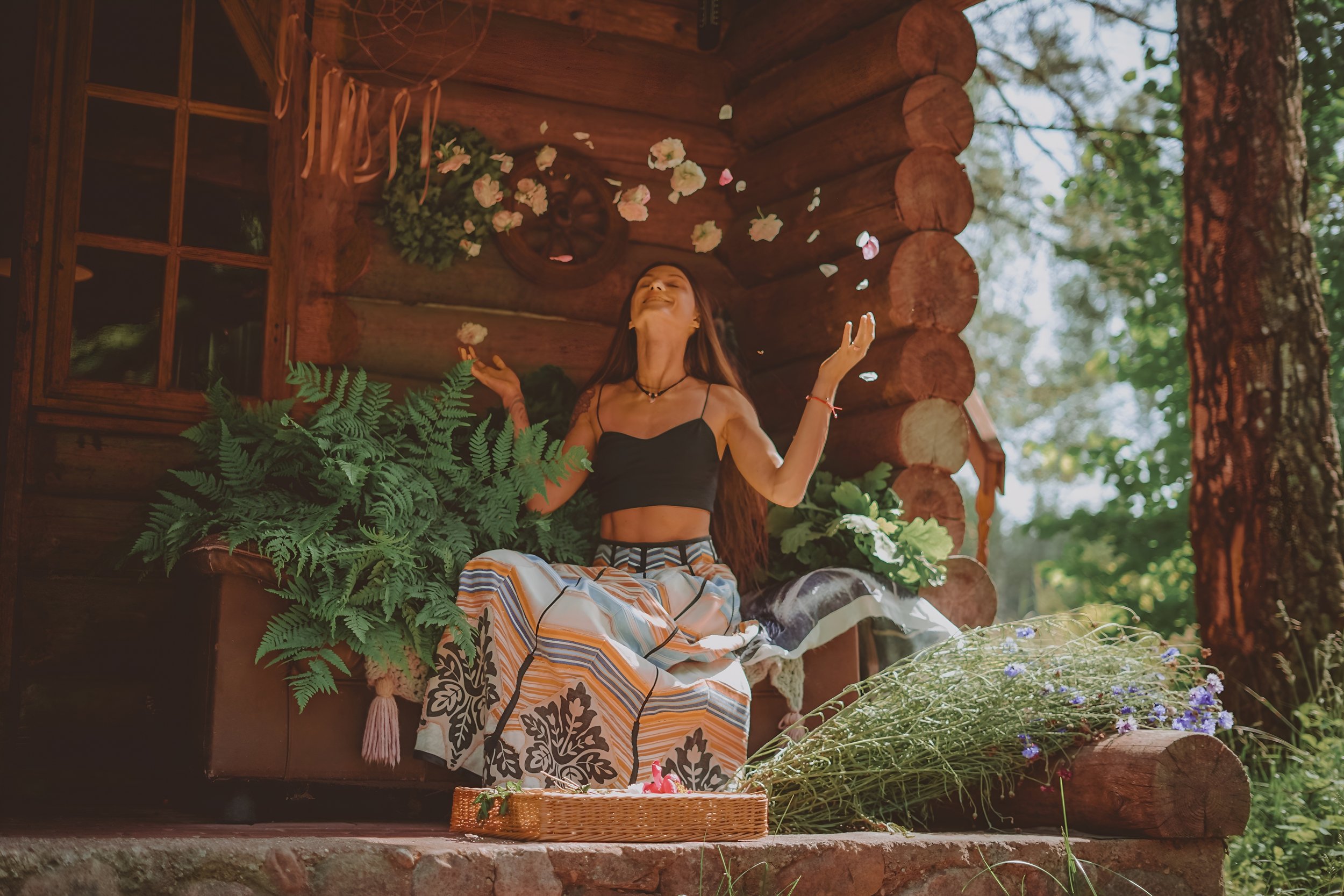 A woman sitting on a wooden porch of a rustic cabin surrounded by greenery and plants, throwing flower petals into the air with a joyful expression.