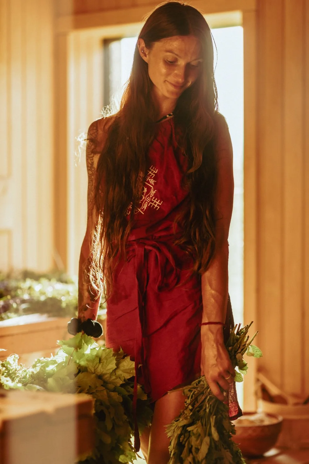 A woman with long brown hair, wearing a red dress with Asian symbols, standing in a wooden interior, holding a bunch of green foliage in her right hand, with warm lighting and windows in the background.