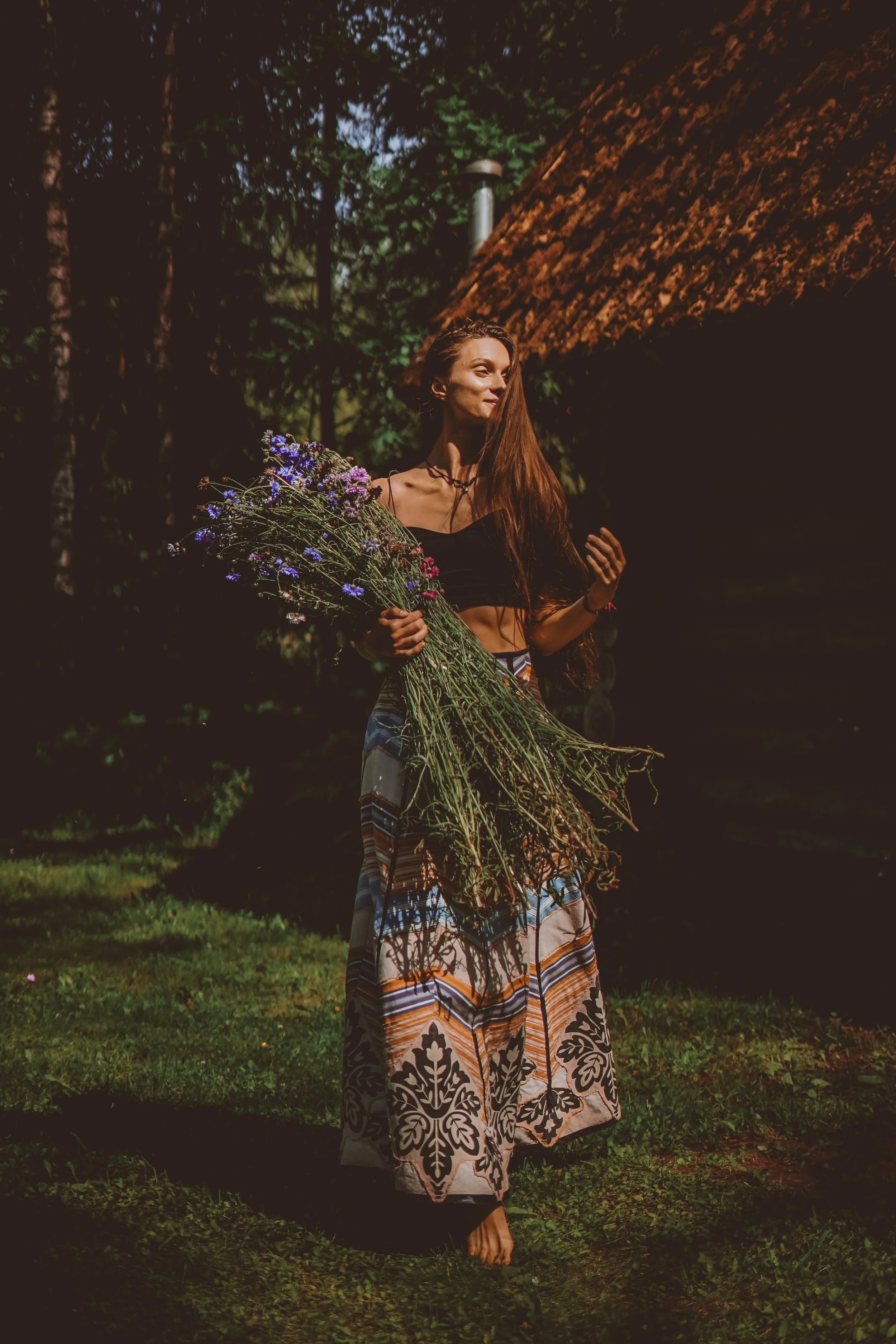 A woman with long brown hair standing outdoors during the daytime, holding a large bouquet of wildflowers, wearing a black crop top and patterned wide-leg pants, with a rustic house with a sloped roof in the background.