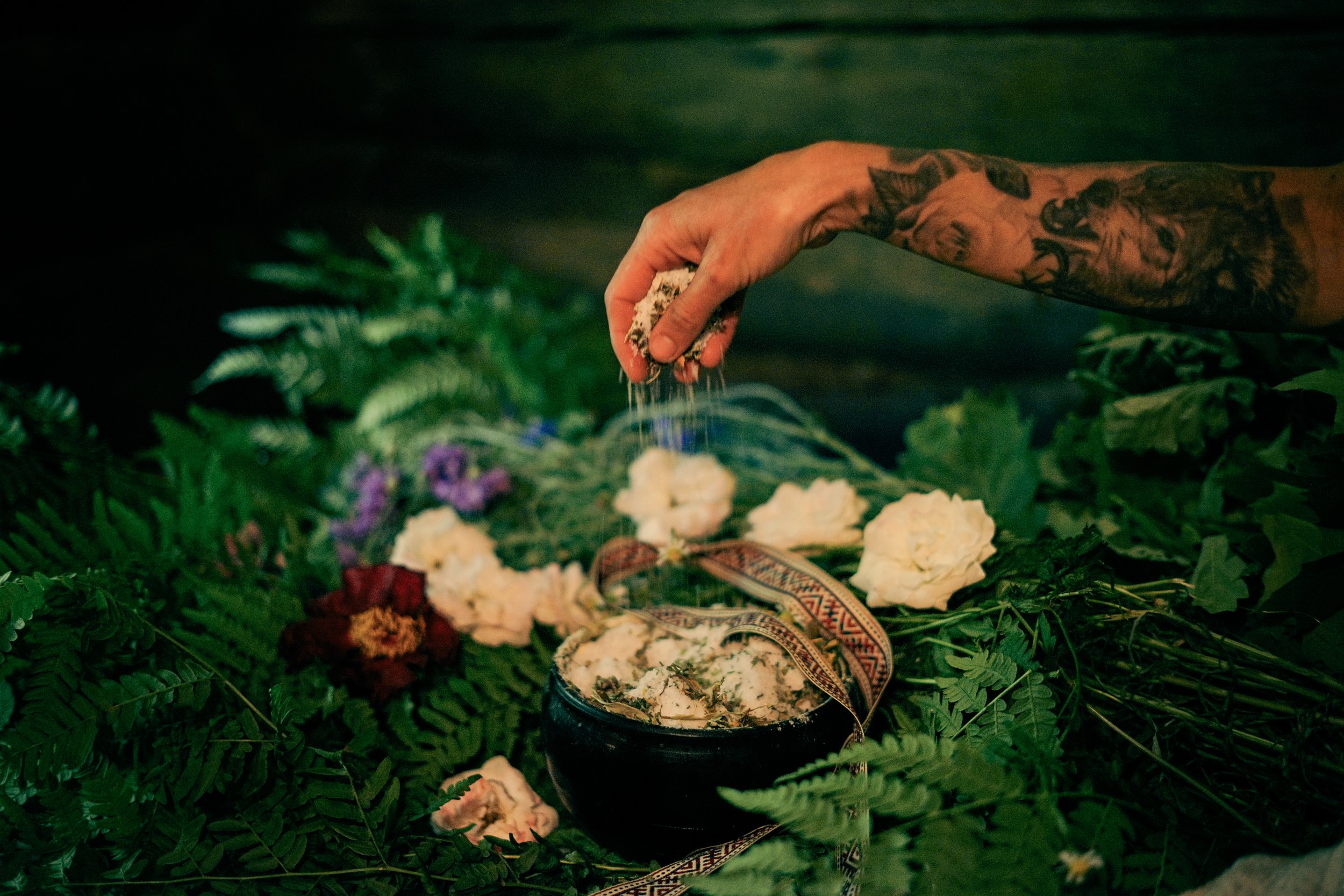 A hand with tattoos sprinkling salt over a decorated floral arrangement, surrounded by ferns and flowers.