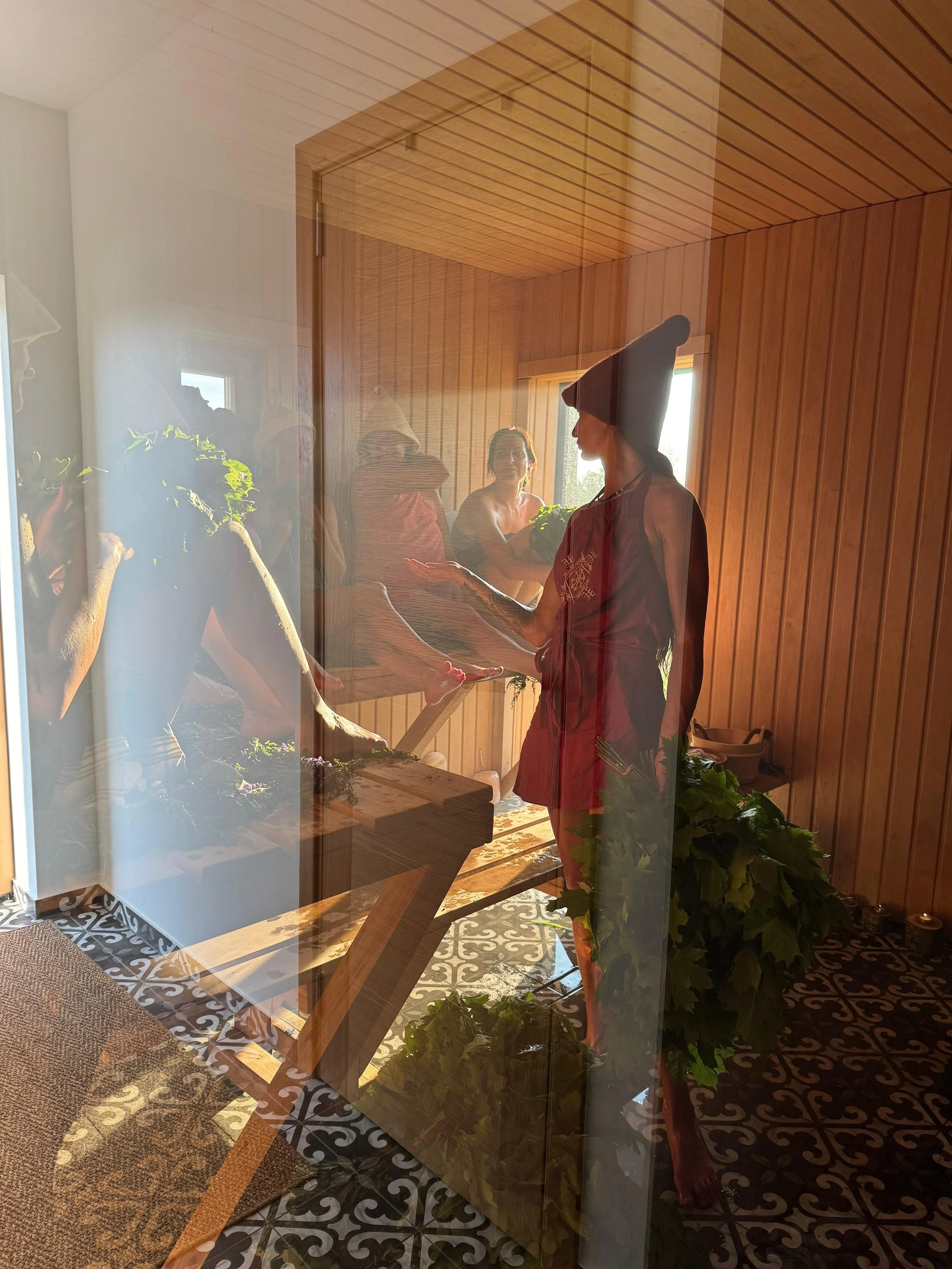 People participating in a sauna session, with one person receiving a treatment, seen through a glass window with reflections.