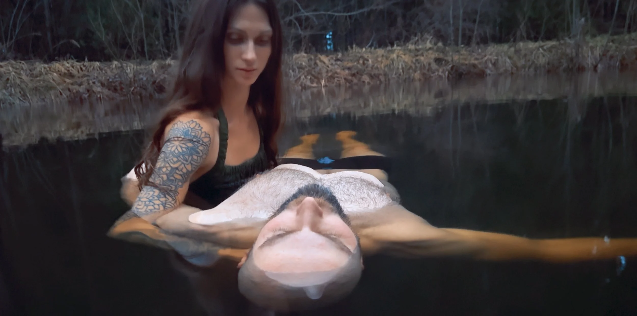 A woman with long dark hair and tattooed arm is in a body of water, supporting a man with a tattooed chest who is floating on his back in a natural outdoor setting.