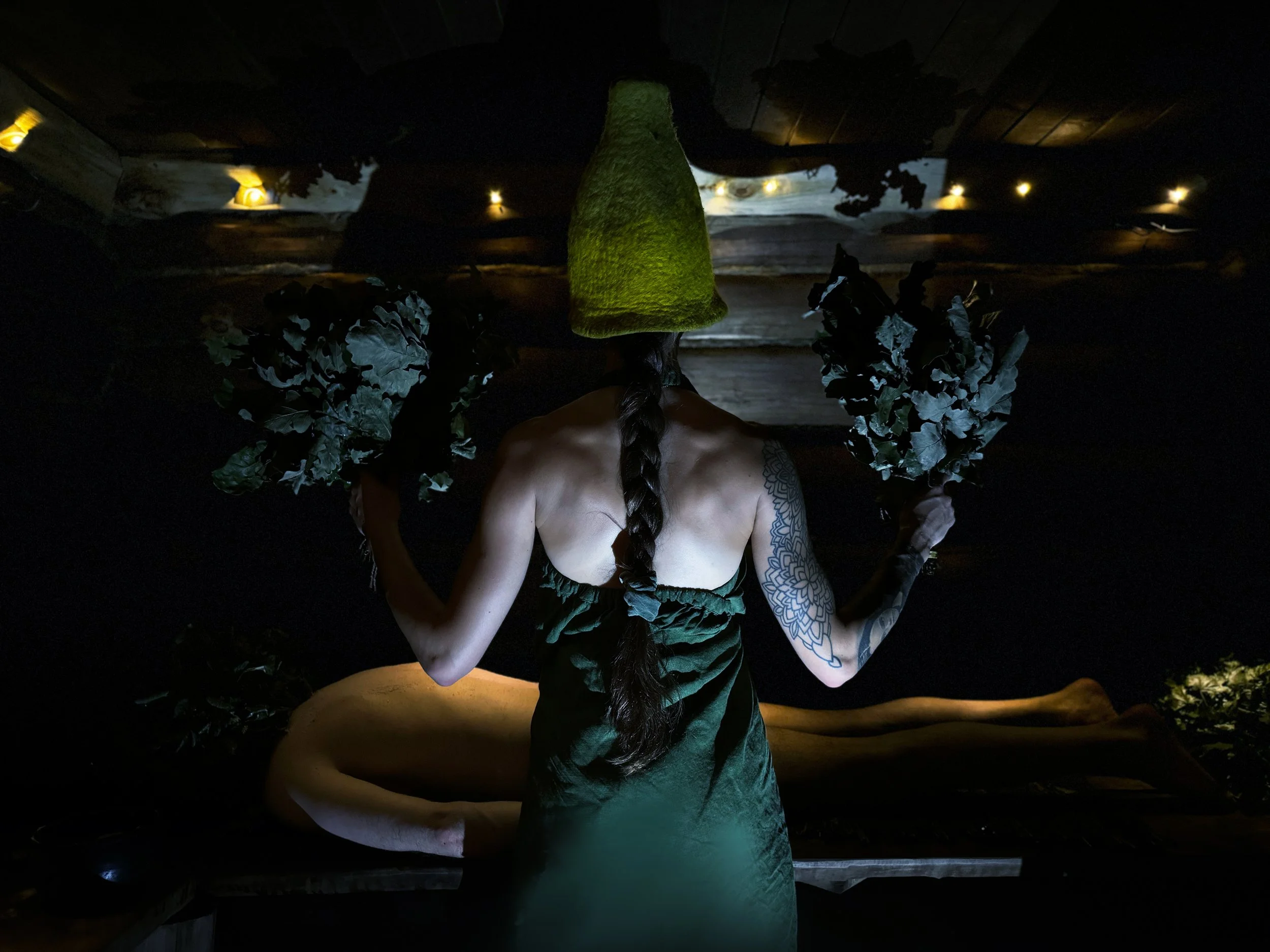A woman with a long braid, tattoos on her arm, wearing a green dress and a yellow felt hat, is sitting with her back facing the camera in a dimly lit room, holding leafy plants in each hand.