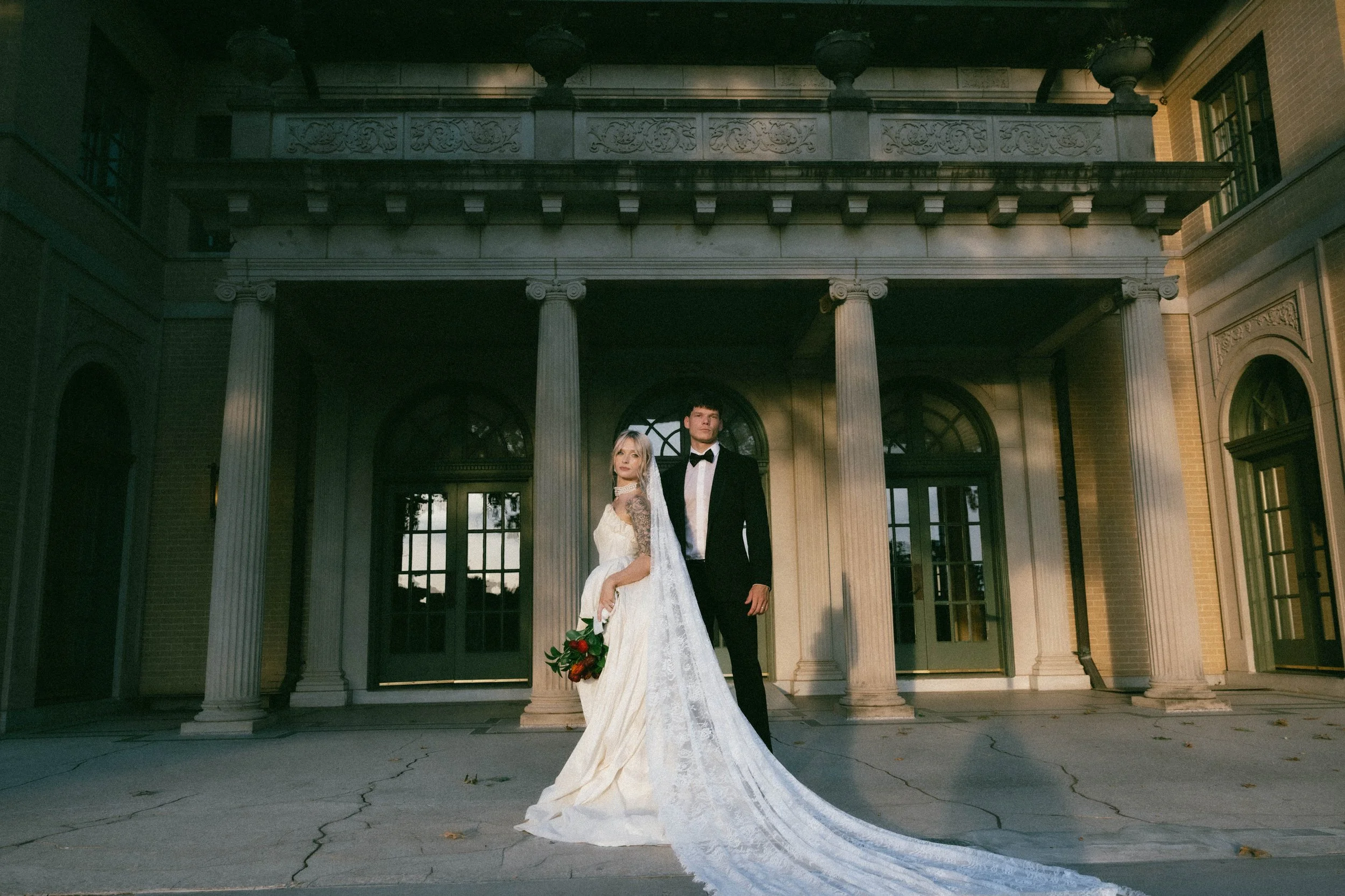 A bride and groom in formal wedding attire pose outdoors framed by European style historic stone architecture in Tulsa, Oklahoma, captured in film inspired , nostalgic style